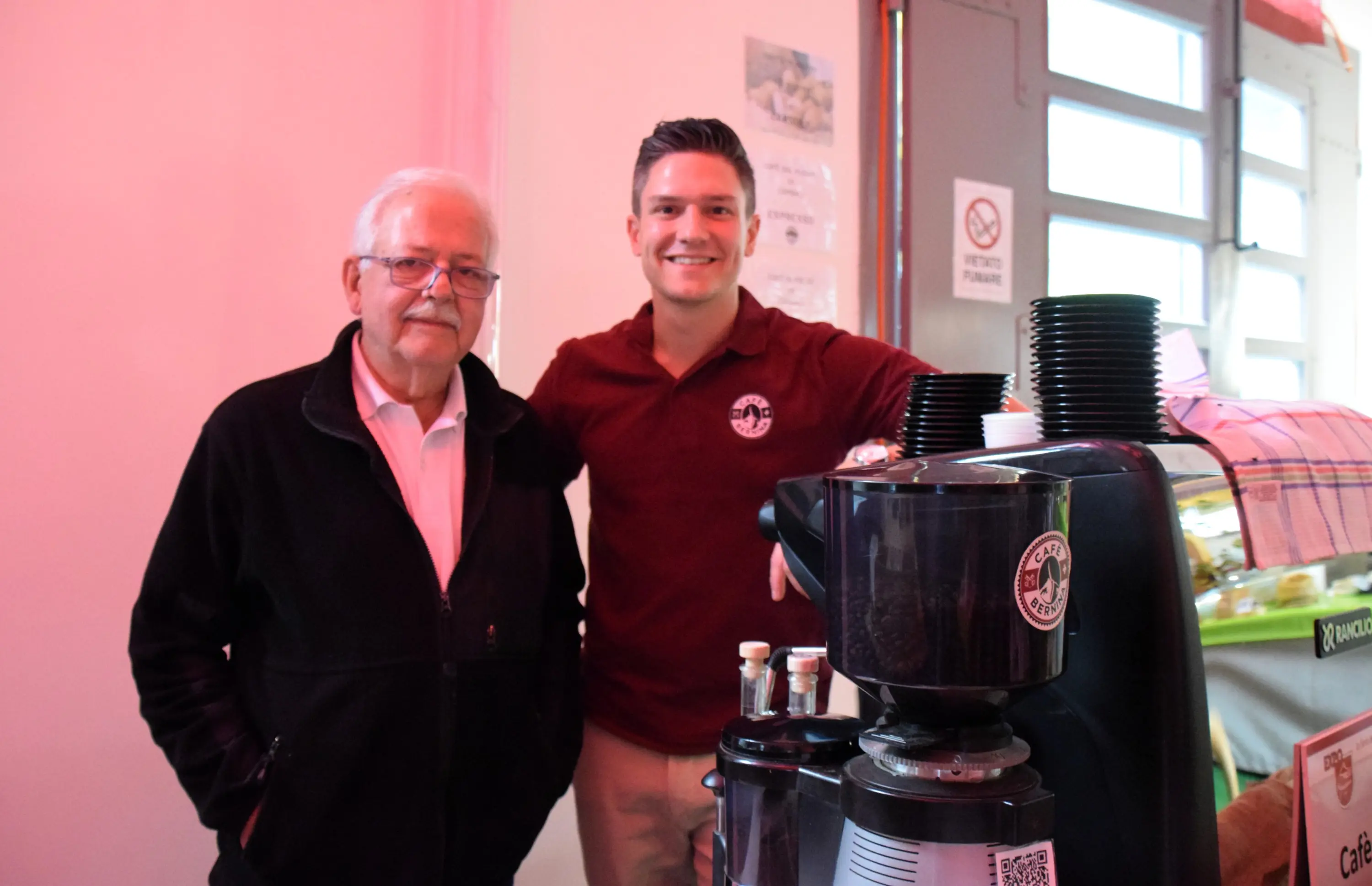 Due uomini sorridenti, uno più anziano con occhiali e un maglione scuro, e un giovane in una polo rossa, si trovano accanto a una macchina del caffè in un ambiente luminoso e colorato.