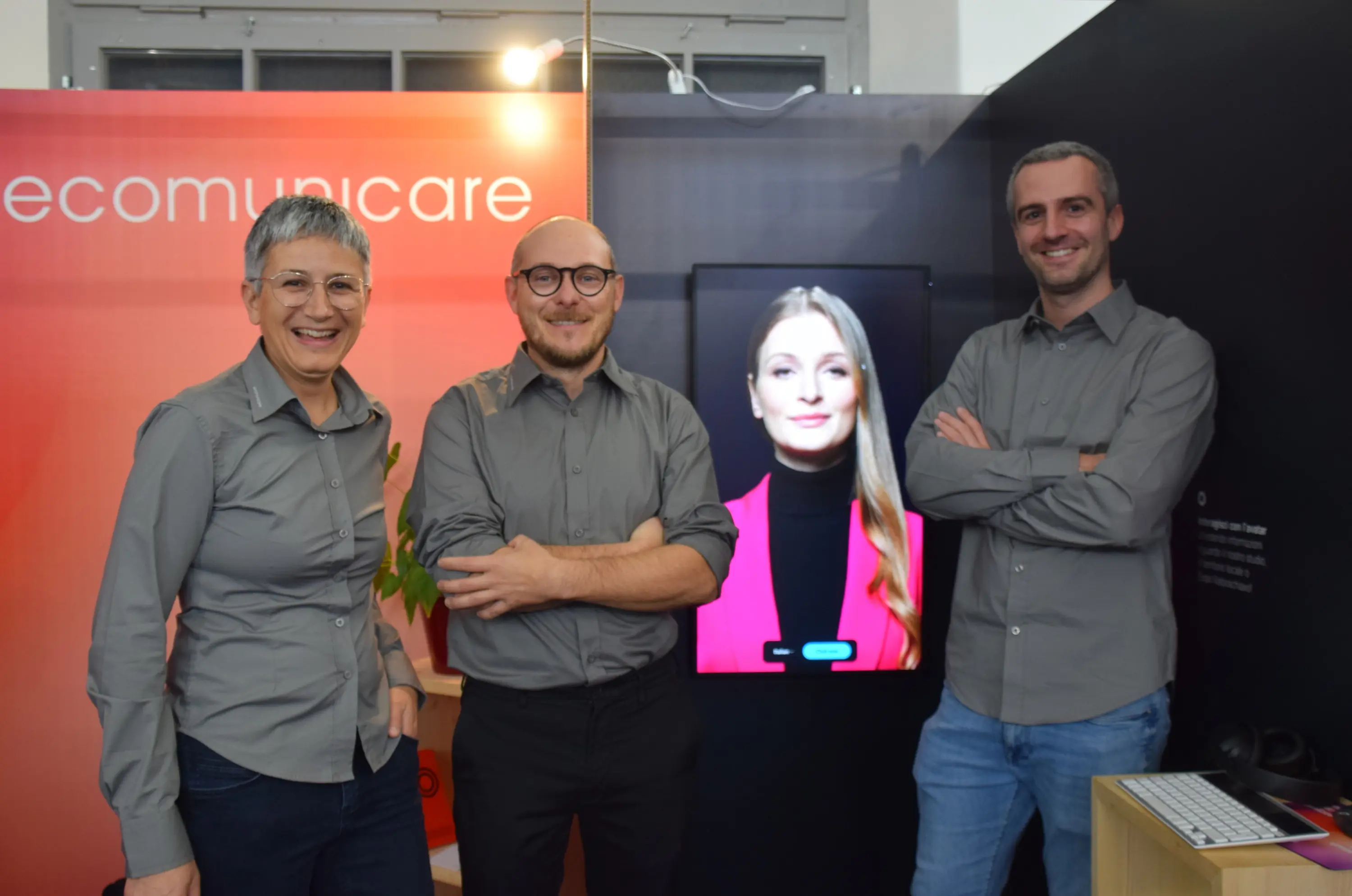 Un gruppo di tre persone sorridenti davanti a uno schermo che mostra un'immagine di una donna. Tutti indossano camicie grigie e si trovano in un ambiente decorato con un tema rosso.