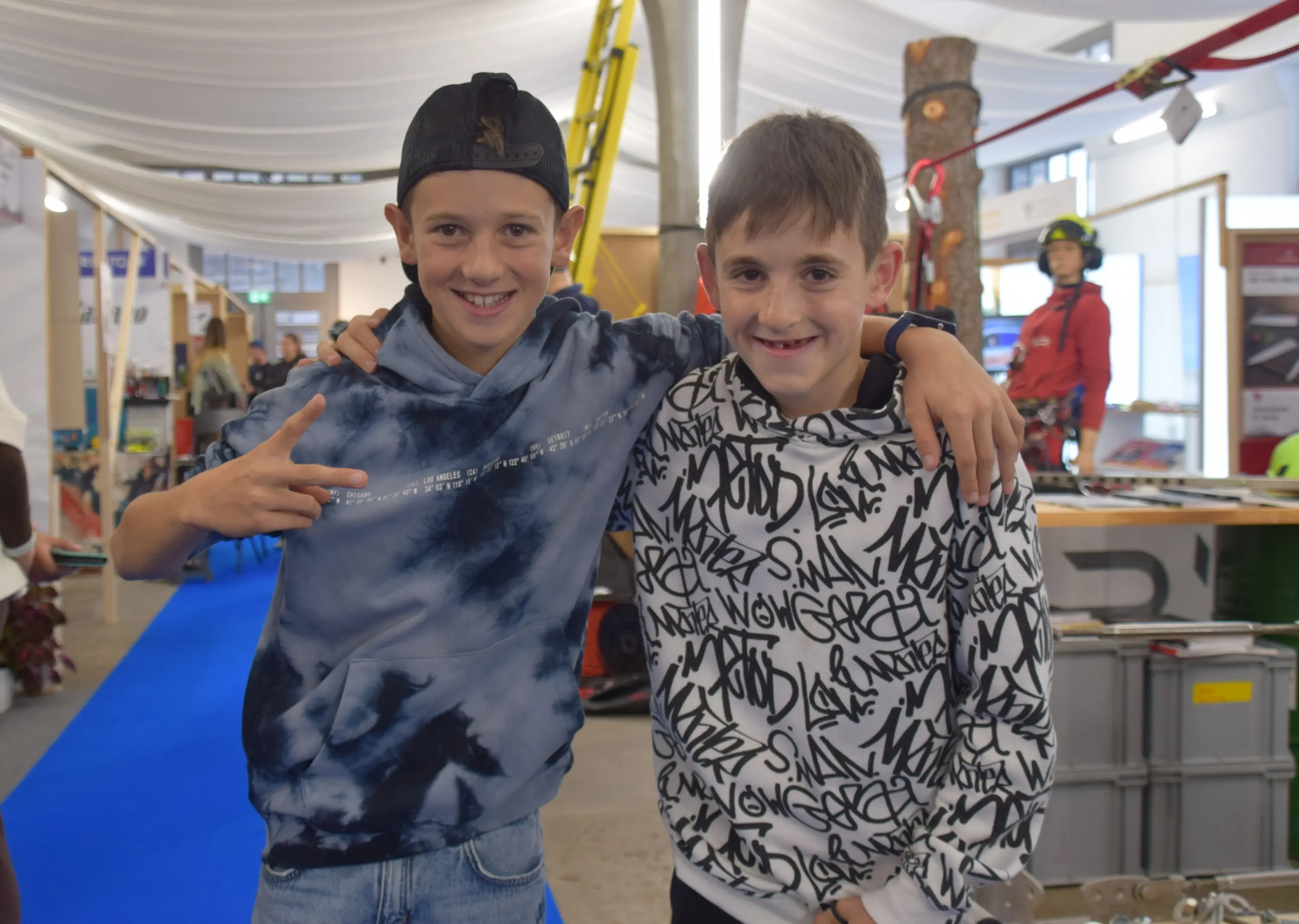 Due ragazzi sorridenti in un ambiente fieristico. Indossano felpe con cappuccio, uno con motivo tie-dye blu e l'altro con un design nero e bianco. Si abbracciano e mostrano un gesto di vittoria.