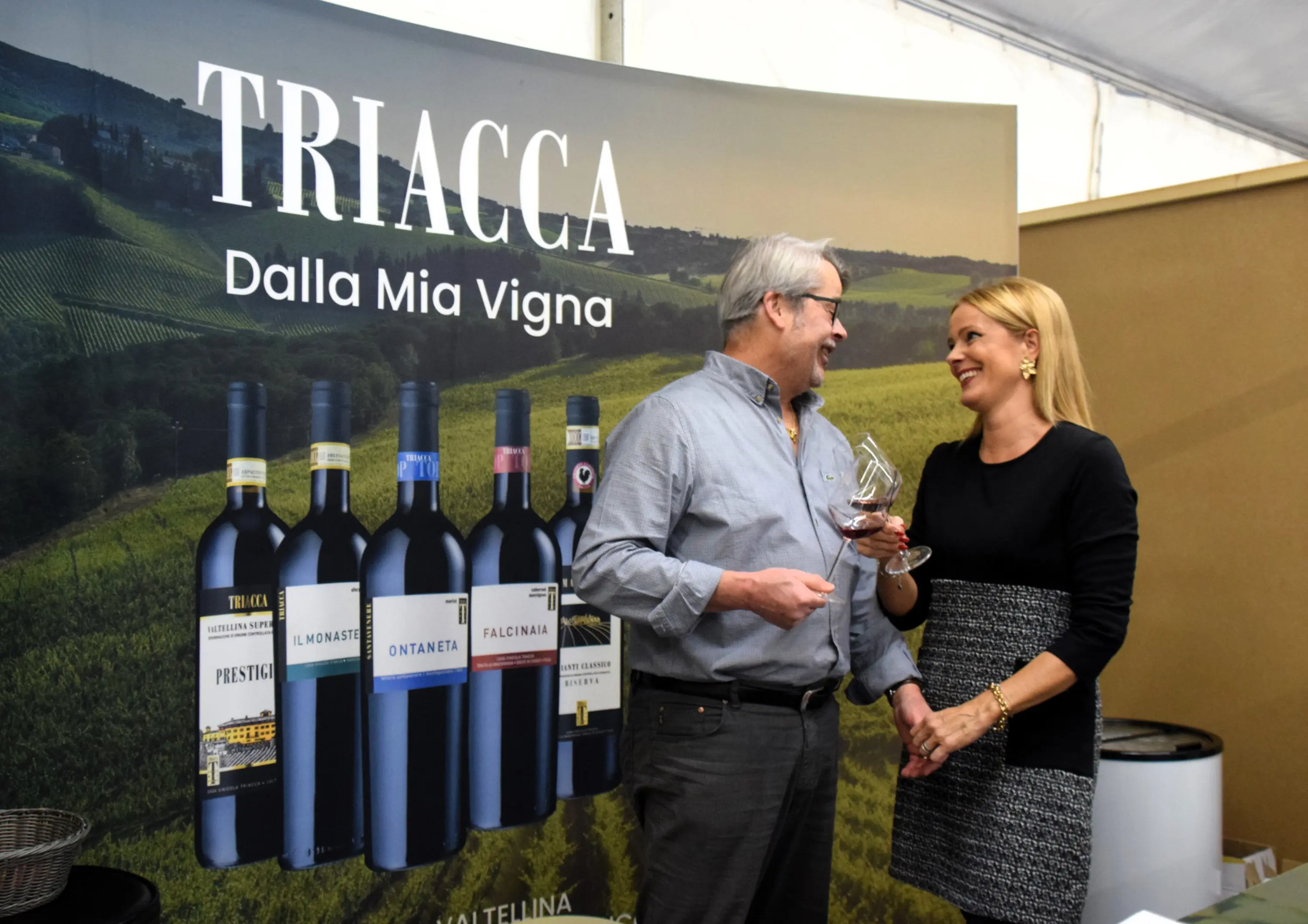 Due persone sorridenti stanno parlando davanti a uno sfondo che mostra diverse bottiglie di vino. L'uomo tiene un bicchiere di vino, mentre la donna sembra interessata alla conversazione.