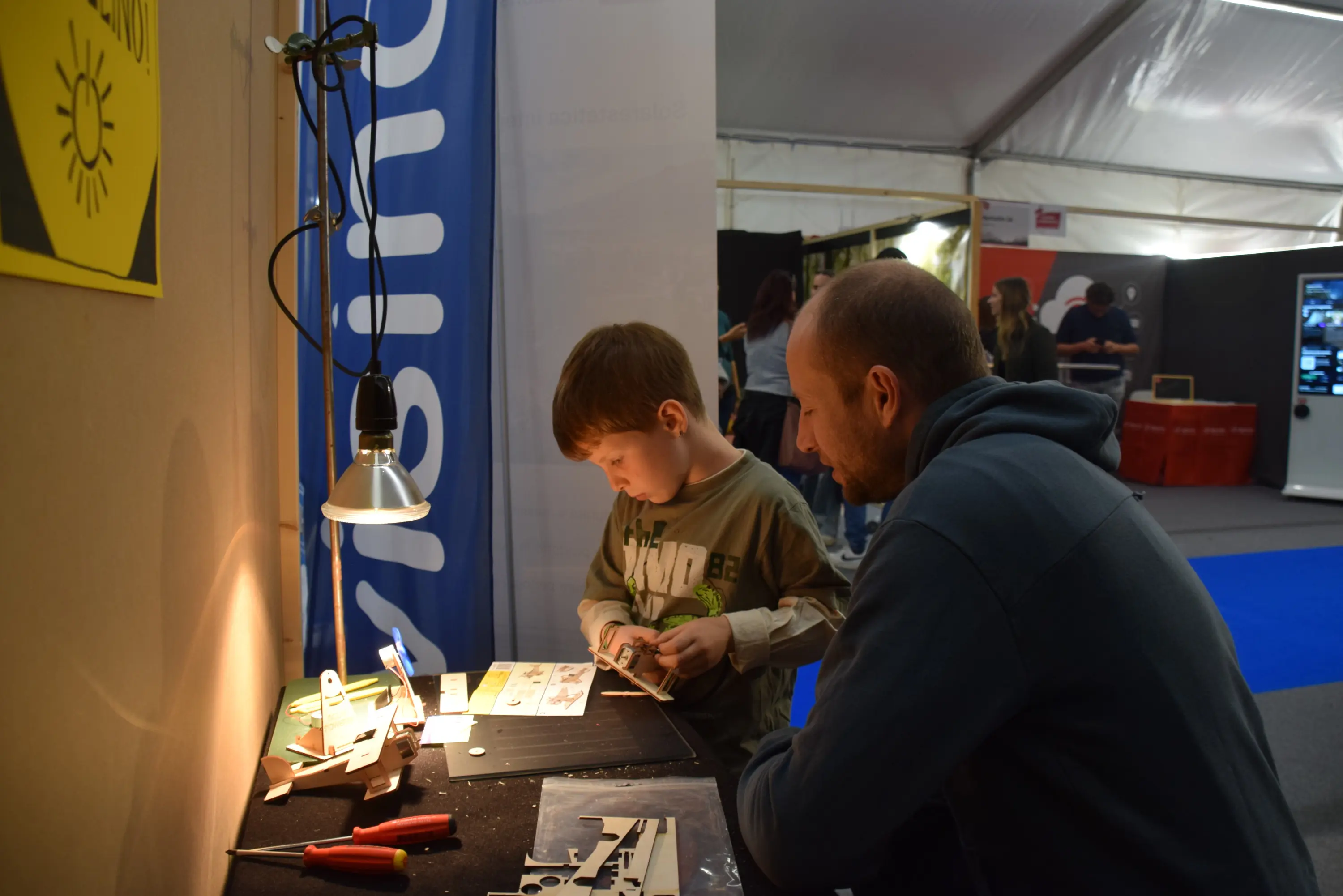 Un bambino e un uomo stanno lavorando insieme a un tavolo, utilizzando strumenti e materiali per un progetto creativo. Il bambino sembra concentrato mentre sistema i pezzi, e l'adulto lo osserva con interesse. Sullo sfondo ci sono altre persone e un ambiente espositivo.