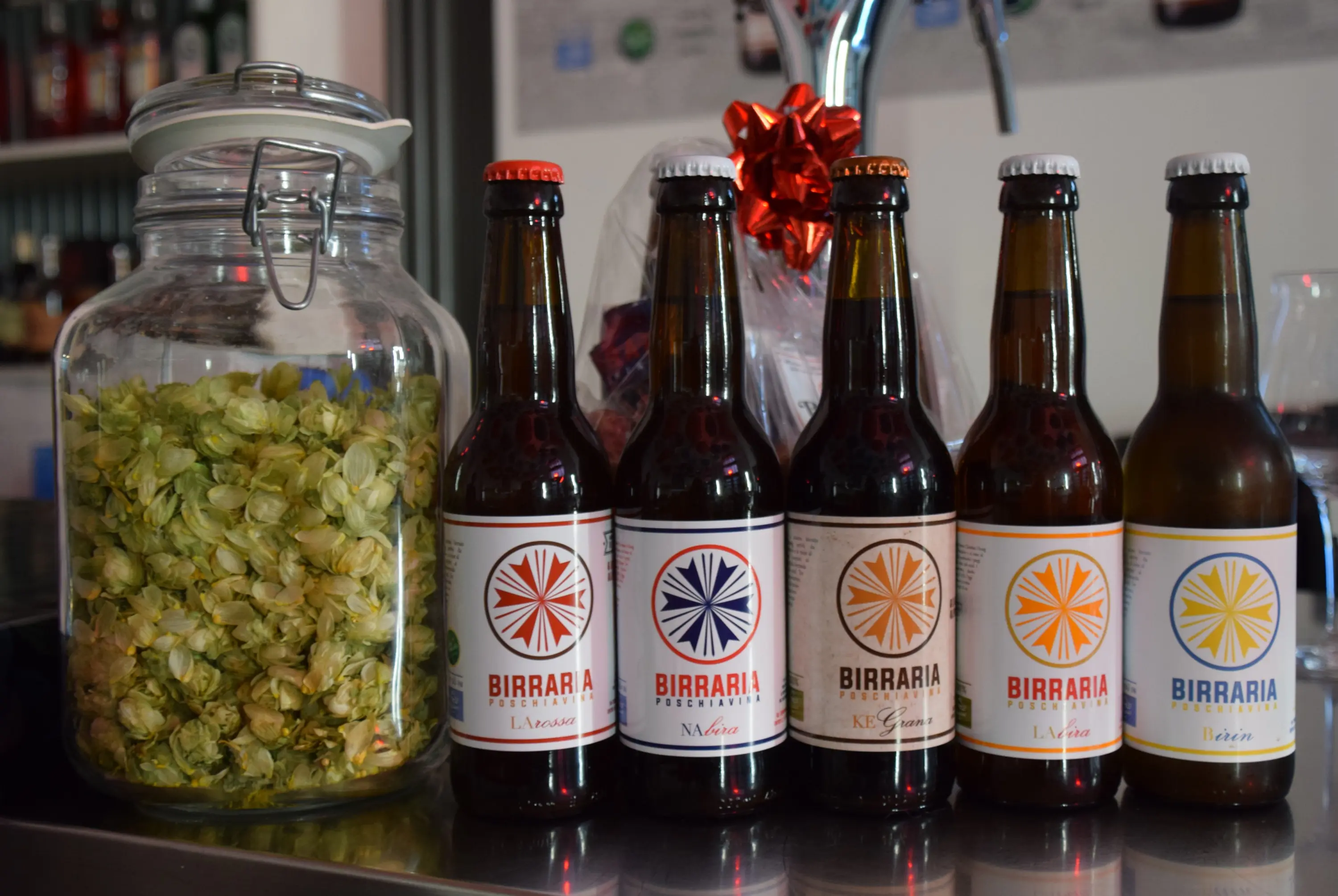 Una selezione di cinque birre artigianali in bottiglia affiancate a un barattolo di luppolo essiccato. I tappi delle bottiglie sono di diversi colori, mentre il barattolo è trasparente e pieno di fiori di lupp