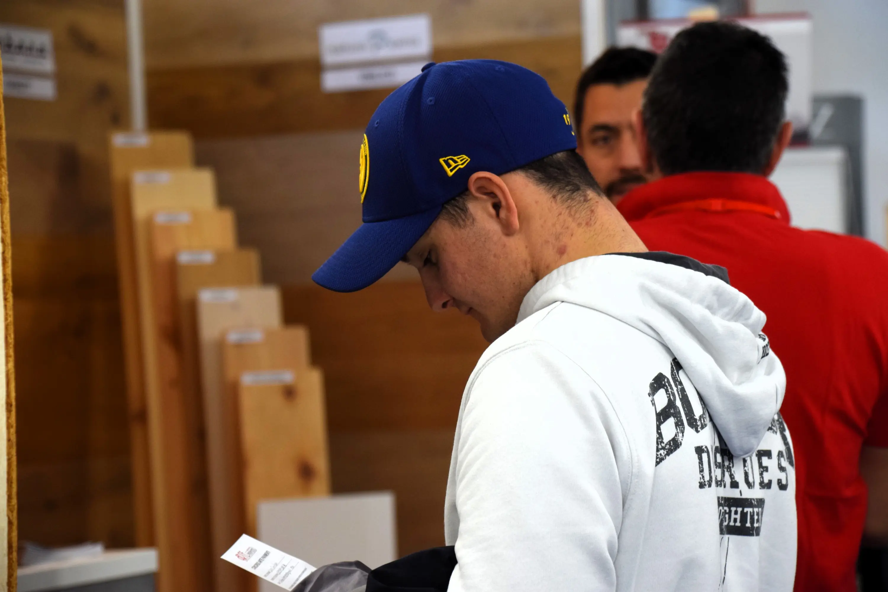 Un uomo con un cappellino blu e una felpa chiara osserva un documento mentre si trova in un ambiente con pannelli di legno e altri due uomini sullo sfondo.