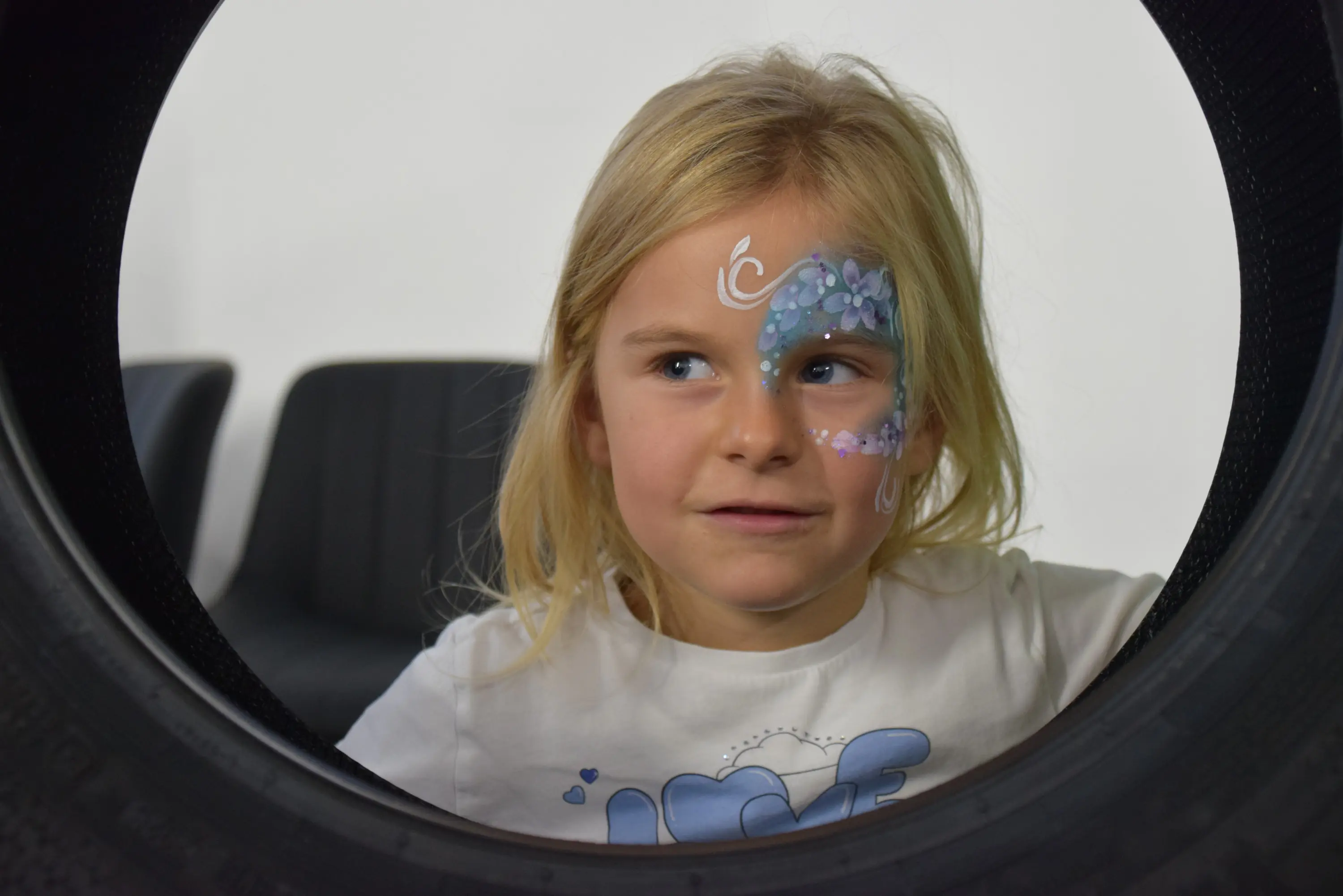 Bambina con capelli biondi e maglietta bianca, sorridente mentre guarda attraverso un pneumatico. Ha un dipinto facciale colorato su una guancia. Sullo sfondo si intravedono delle sedie nere.
