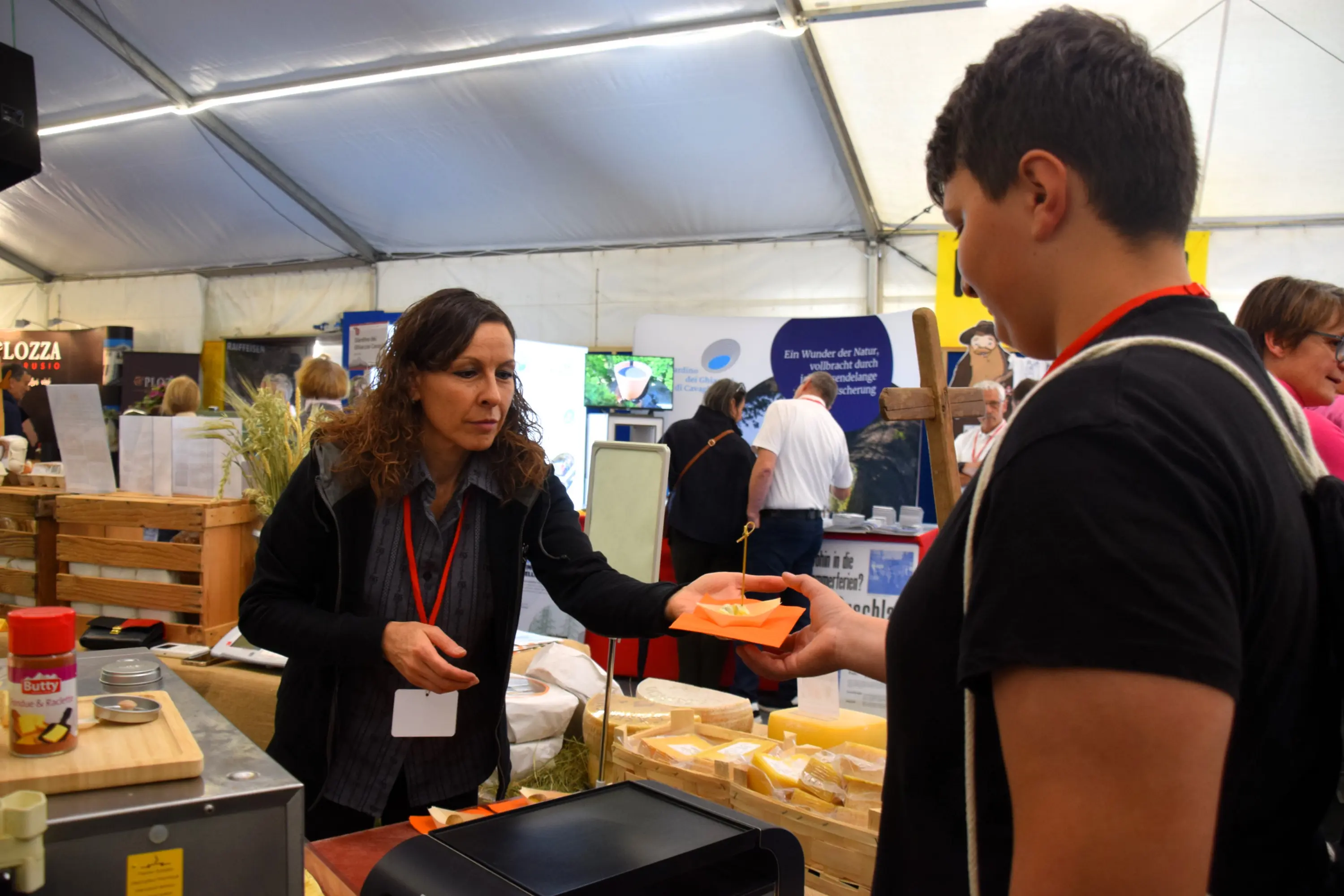 Una donna sta condividendo un campione di formaggio con un ragazzo in una fiera. Intorno, ci sono vari espositori e visitatori. La donna indossa una maglietta scura e un distintivo, mentre il ragazzo tiene un sacchetto sulla spalla
