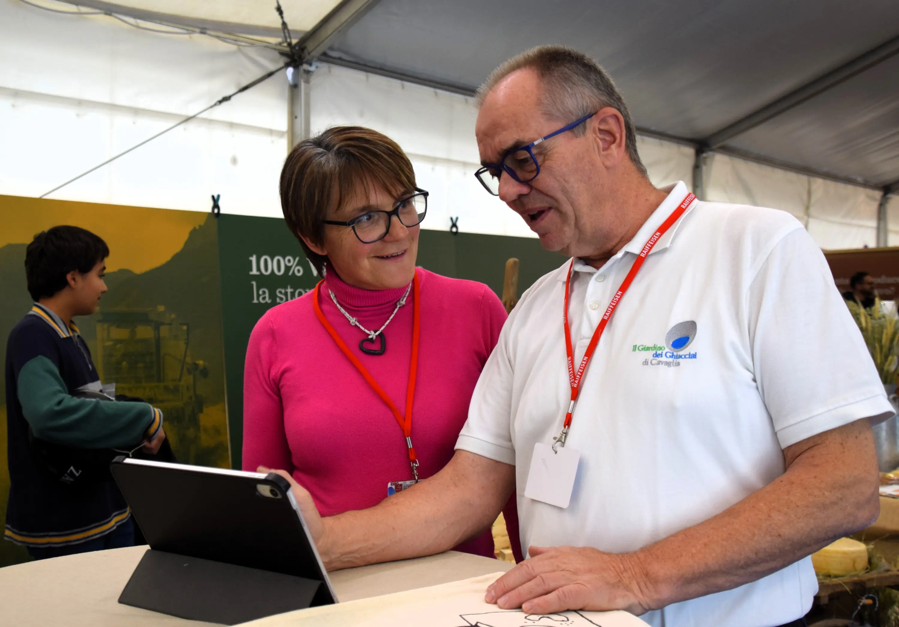 Due persone stanno interagendo davanti a un tablet, con un'atmosfera conviviale. Un uomo, con occhiali e una maglietta bianca, spiega qualcosa all'altra persona, una donna con occhiali e una maglia rosa. Sullo sfondo, si intr