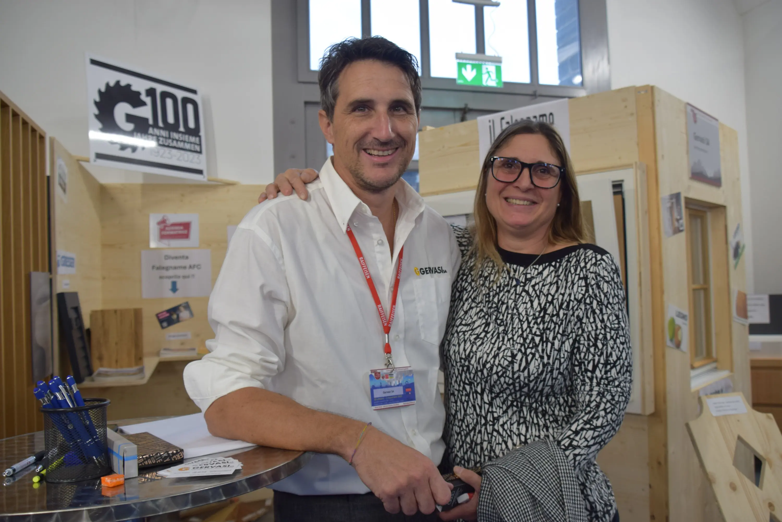 Due persone sorridenti posano insieme in un ambiente di fiera. Un uomo in camicia bianca e cravatta rossa è accanto a una donna con occhiali e maglione a fantasia. Sullo sfondo ci sono espositori di legno e materiale promozionale