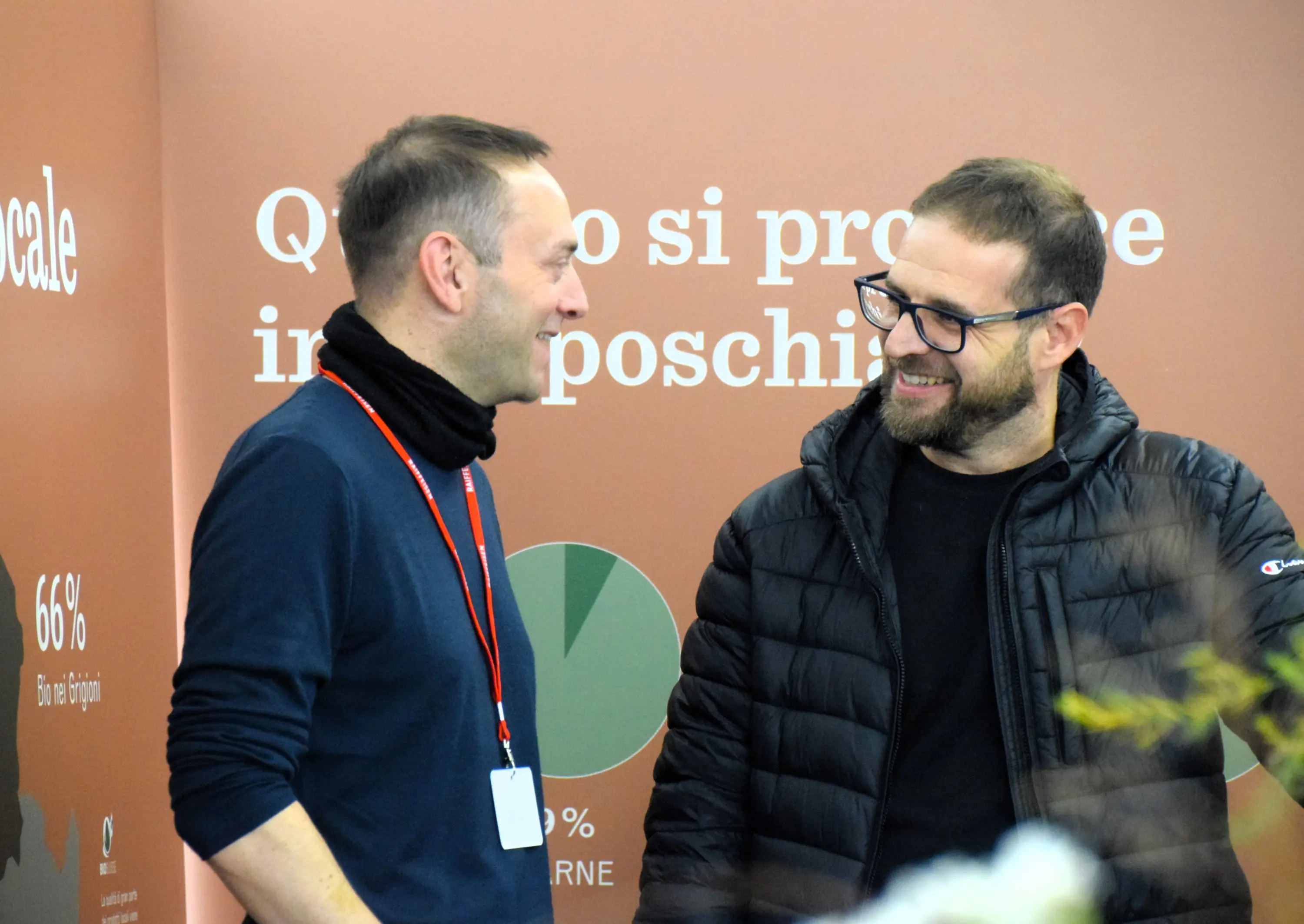 Due uomini sorridono e chiacchierano in un ambiente informale. Sullo sfondo, un pannello informativo con statistiche e testi. Entrambi indossano abbigliamento casual.