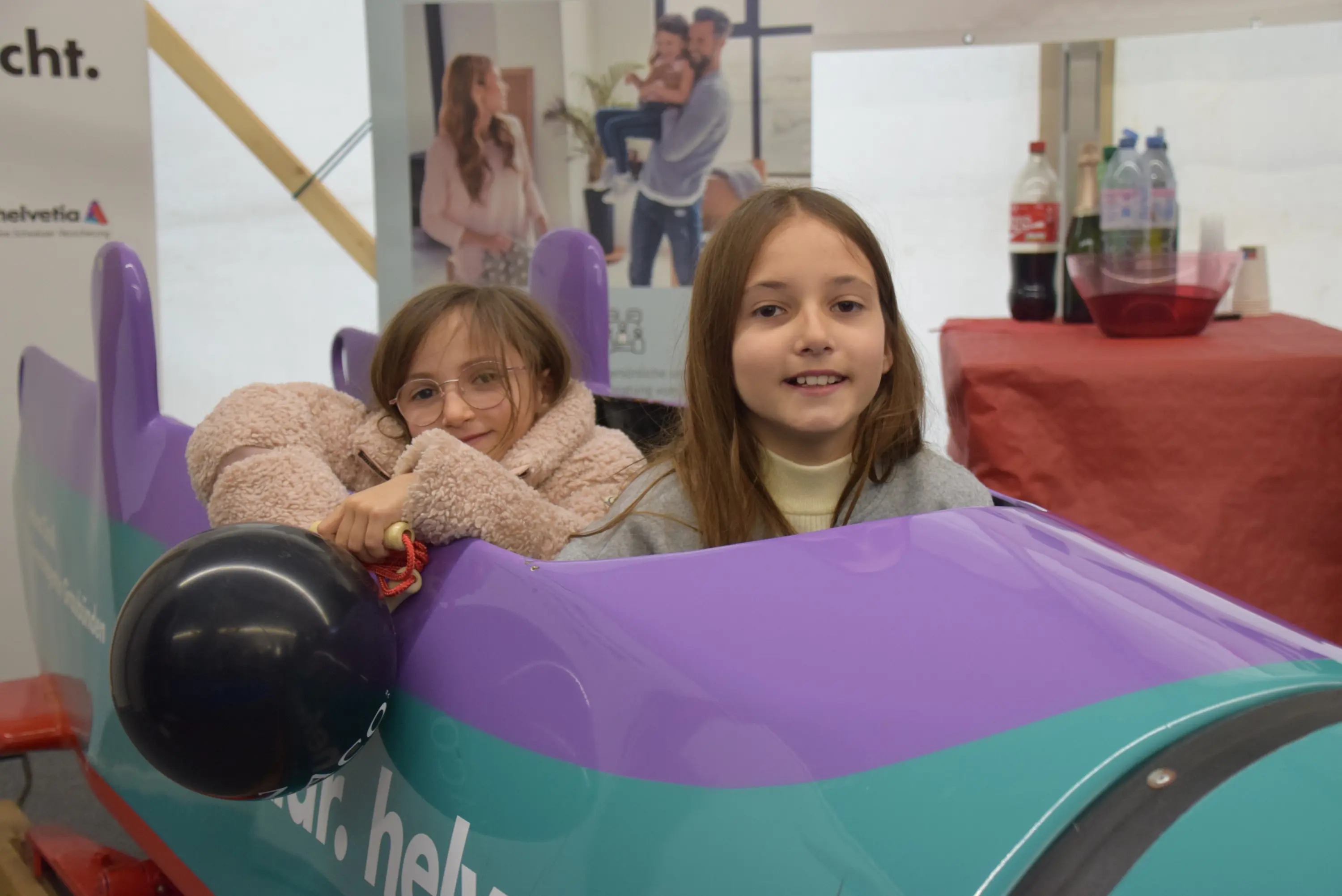 Due bambine sorridenti sedute in un'auto giocattolo colorata, una con occhiali e un palloncino nero. Sullo sfondo, ci sono bottiglie di plastica e un poster.