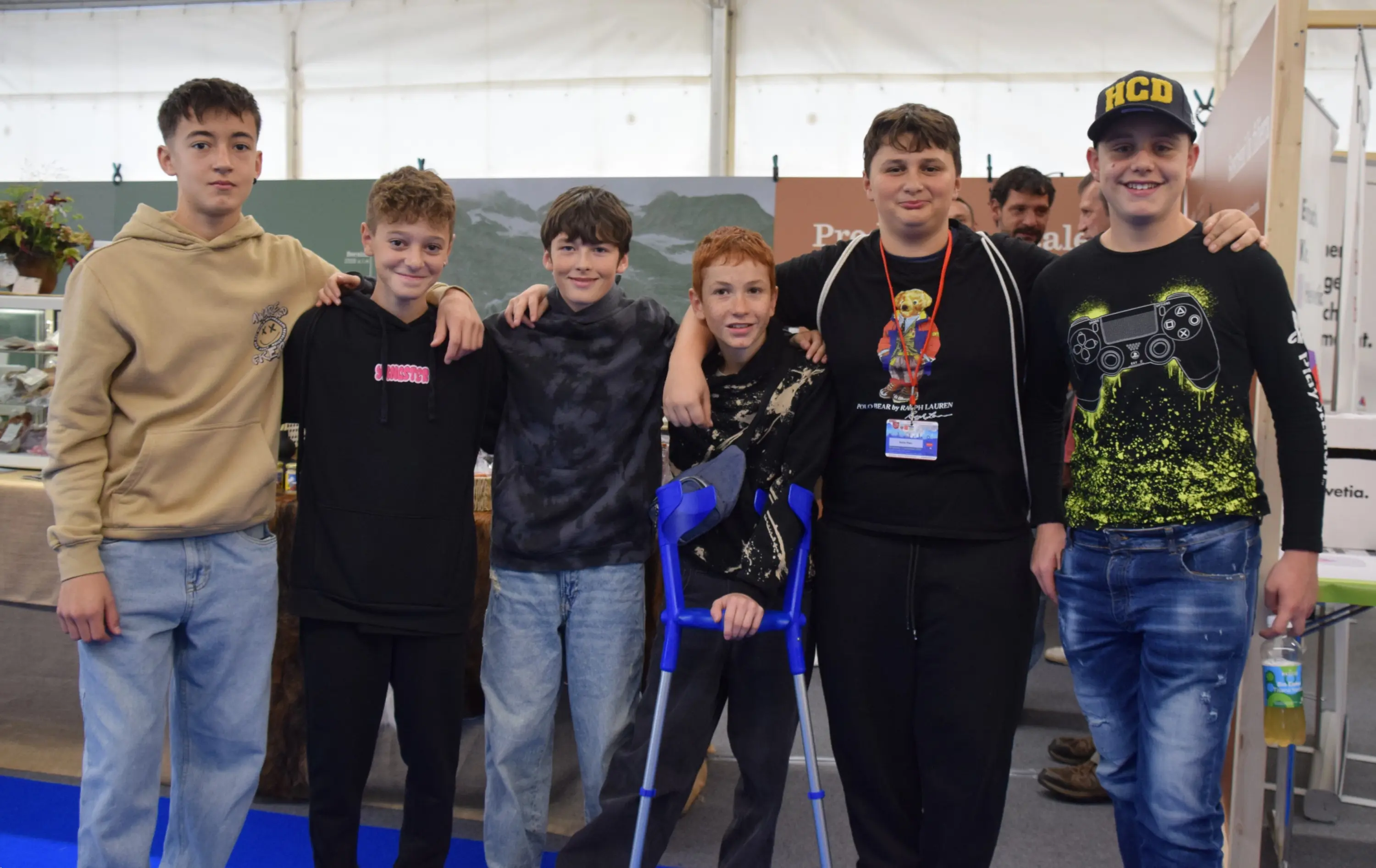 Gruppo di sei ragazzi sorridenti, alcuni indossano felpe e T-shirt casual, in una fiera. Due ragazzi hanno le braccia attorno alle spalle degli altri. Uno di loro utilizza delle stampelle. Sullo sfondo si intravedono oggetti espositivi