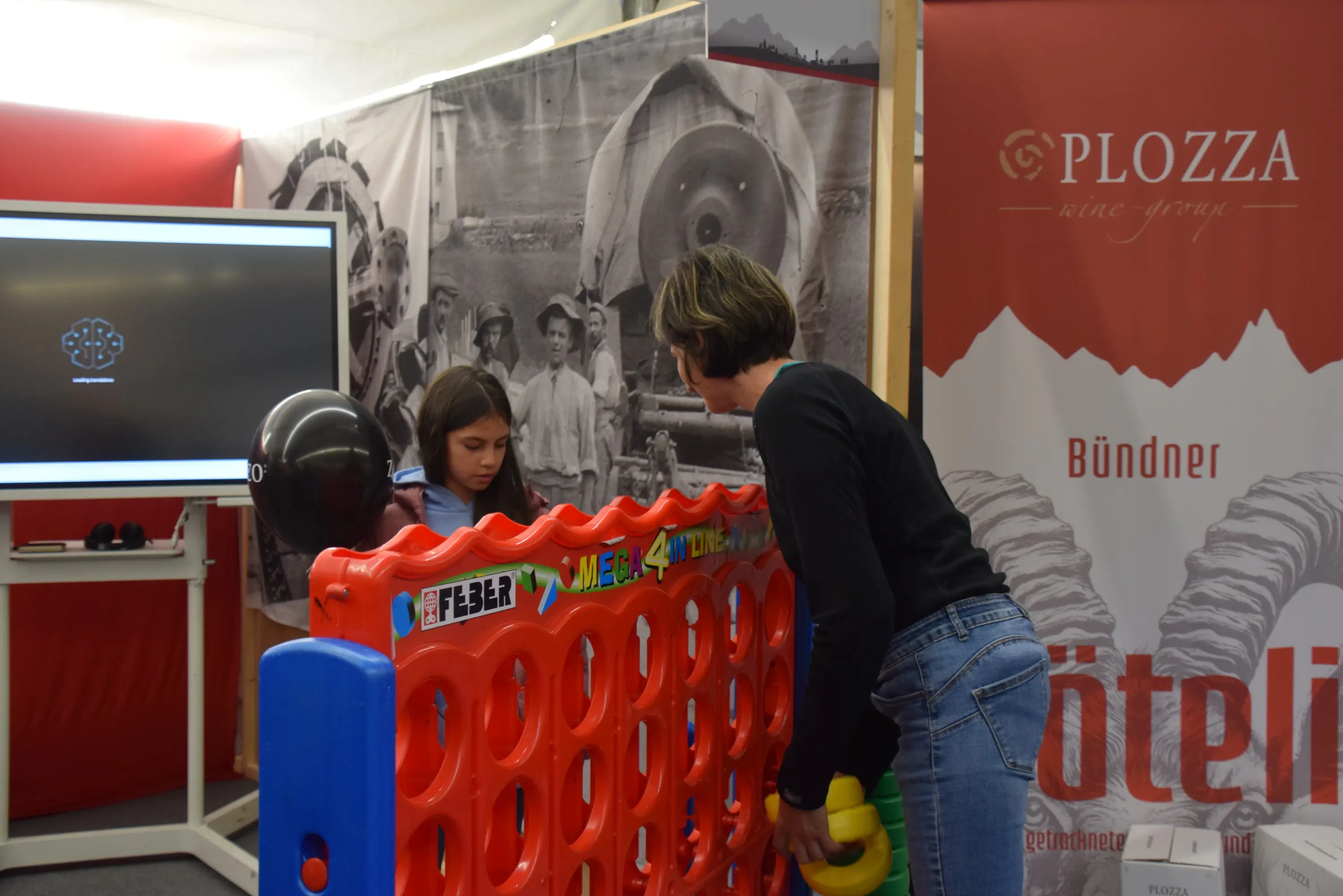 Una ragazza gioca a un gioco di connect four con l'aiuto di un adulto in un ambiente espositivo. Sullo sfondo, ci sono immagini in bianco e nero di persone e una grande affermazione di un marchio.