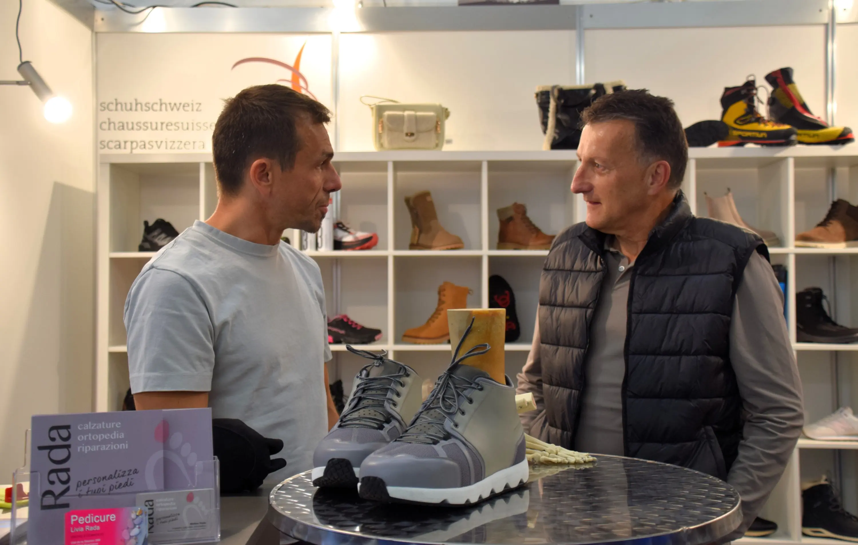 Due uomini stanno conversando a un evento, con una coppia di scarpe grigie al centro del tavolo. Sullo sfondo, scaffali mostrano diversi modelli di calzature. Uno degli uomini indossa una maglia chiara e l'altro una giacca sc