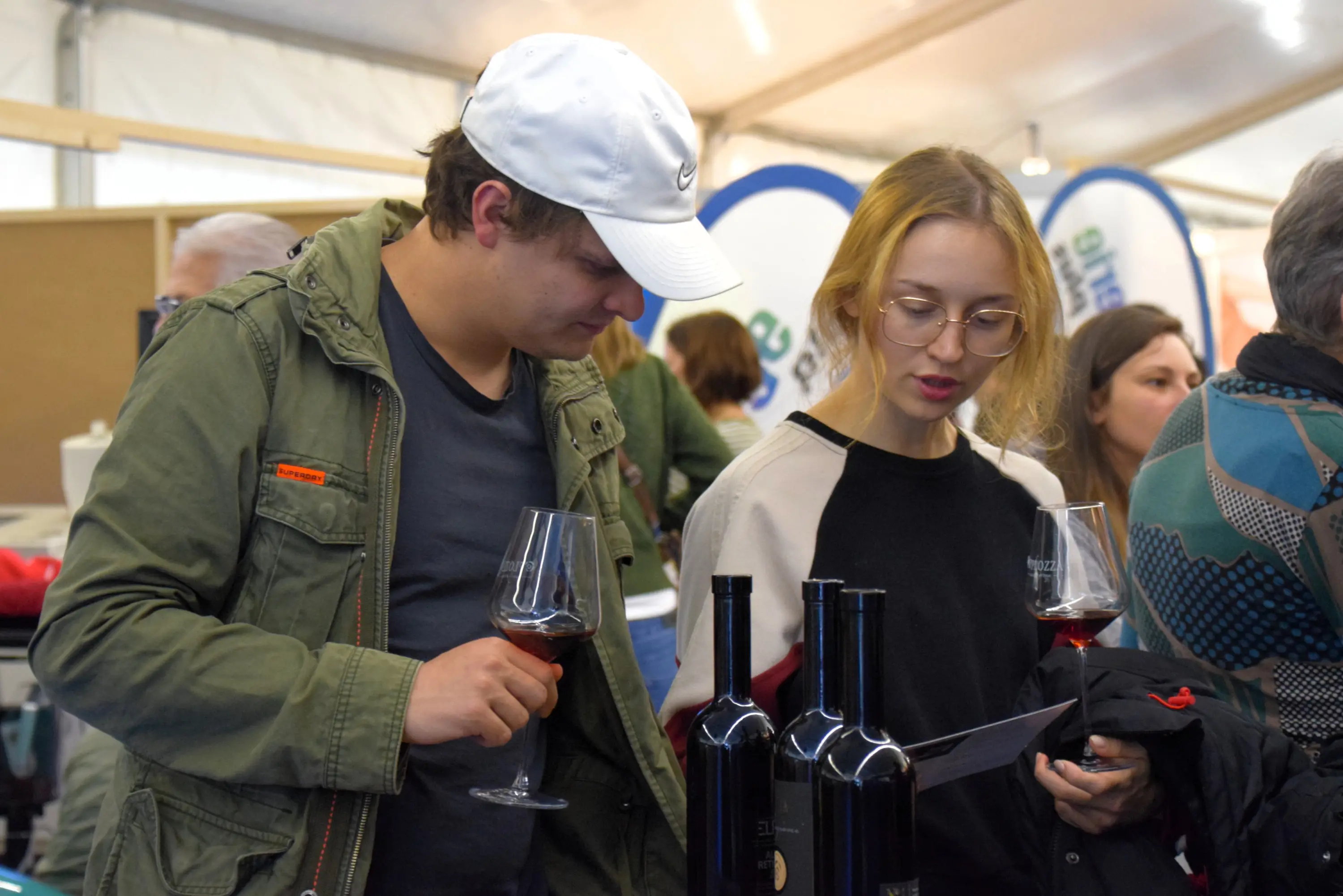 Due giovani, un ragazzo e una ragazza, osservano attentamente un menu mentre tengono in mano dei bicchieri di vino rosso. Sul tavolo ci sono alcune bottiglie di vino. Siamo all'interno di una fiera allestita in una tenda.