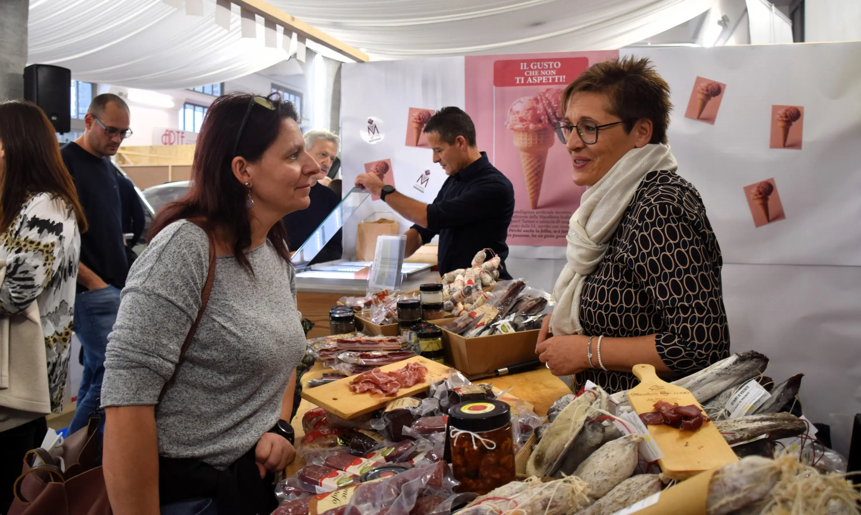 Due donne conversano davanti a un banco di salumi e prodotti gastronomici, mentre sullo sfondo si vede una fiera con altre persone impegnate. Un cartello promozionale è visibile dietro di loro.