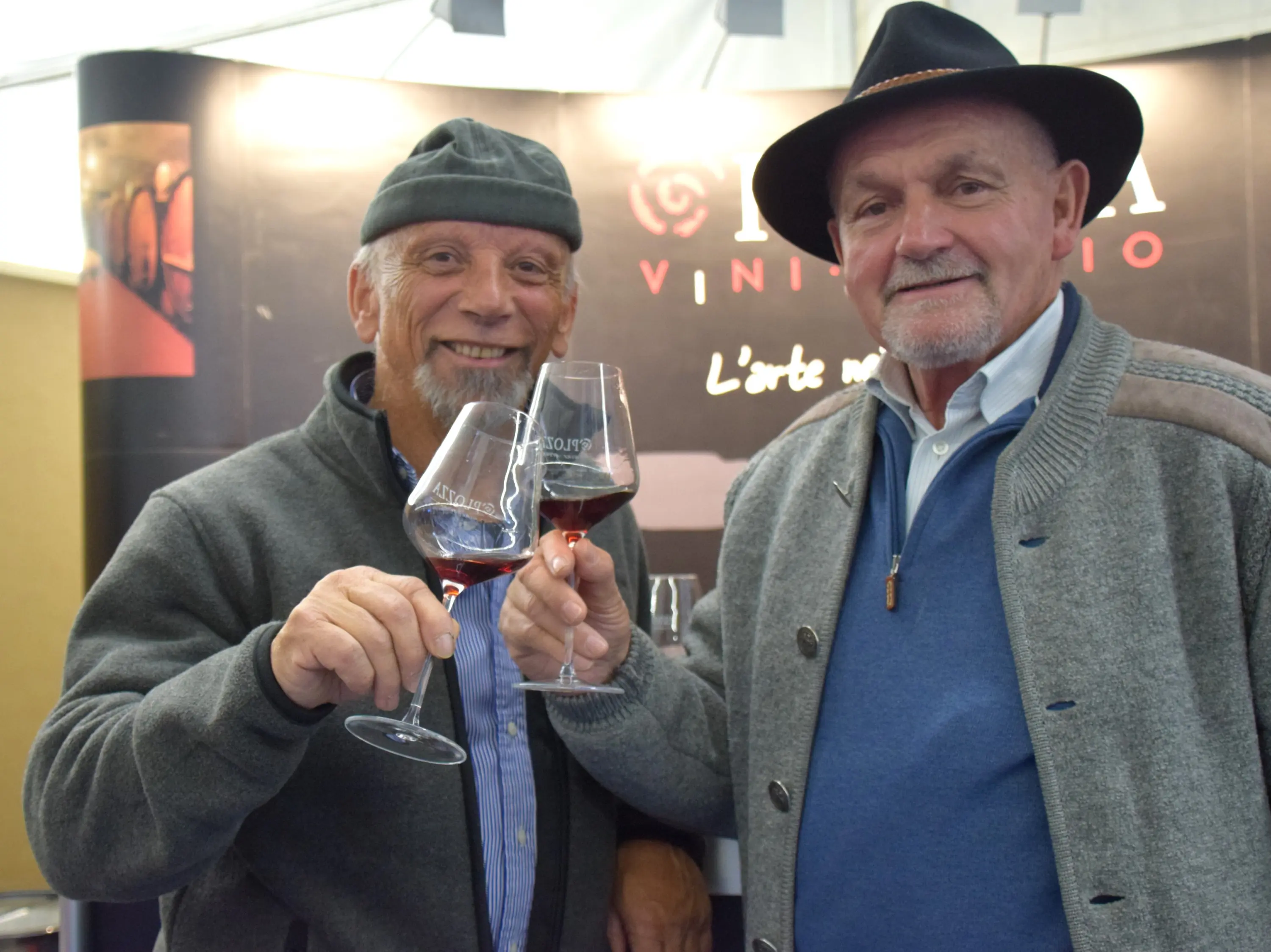 Due uomini sorridenti alzano i calici di vino, mentre si trovano davanti a uno sfondo con il logo di un'azienda vinicola. Indossano cappelli e maglioni di lana.
