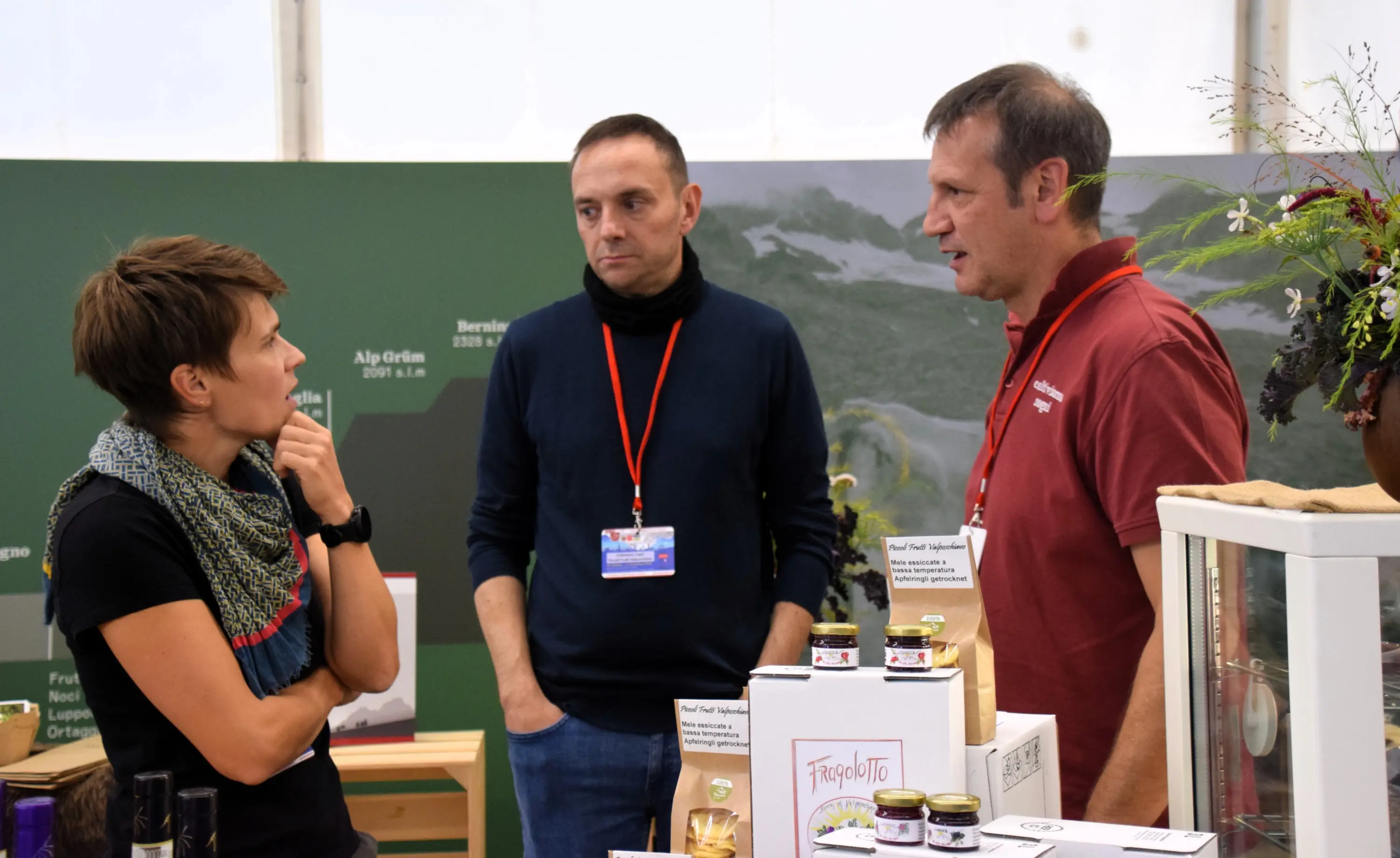 Tre persone conversano in un ambiente espositivo. Due uomini, uno con una maglietta rossa e l'altro con una maglietta nera, ascoltano attentamente una donna che indossa una sciarpa. Sul tavolo ci sono vari prodotti alimentari e decorazioni