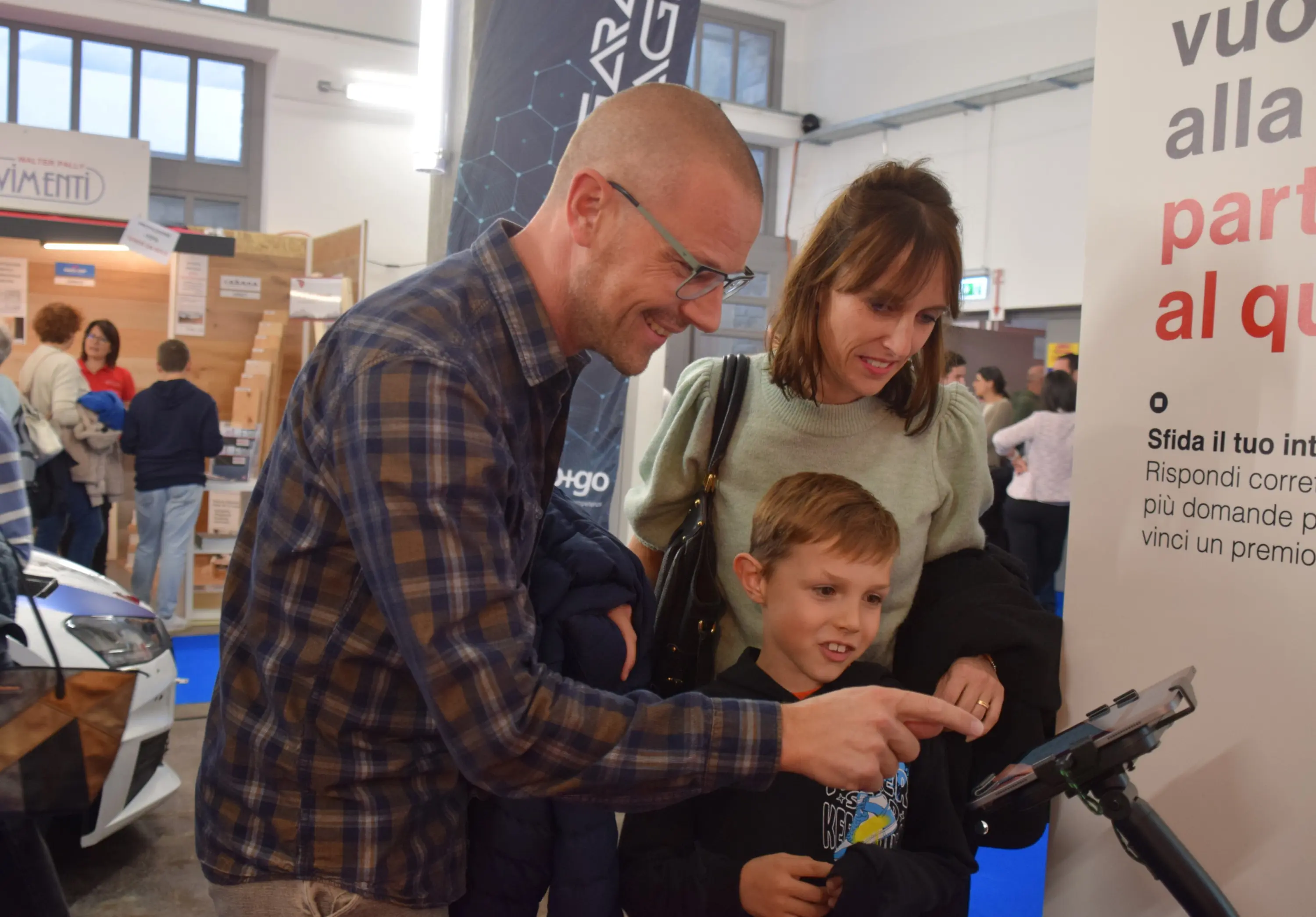 Una famiglia interagisce con un display informatico a un evento, osservando attentamente. L'uomo indossa una camicia a quadri, la donna ha i capelli lunghi e un sorriso, mentre il bambino tocca lo schermo con curiosità. Sullo sfondo si intr