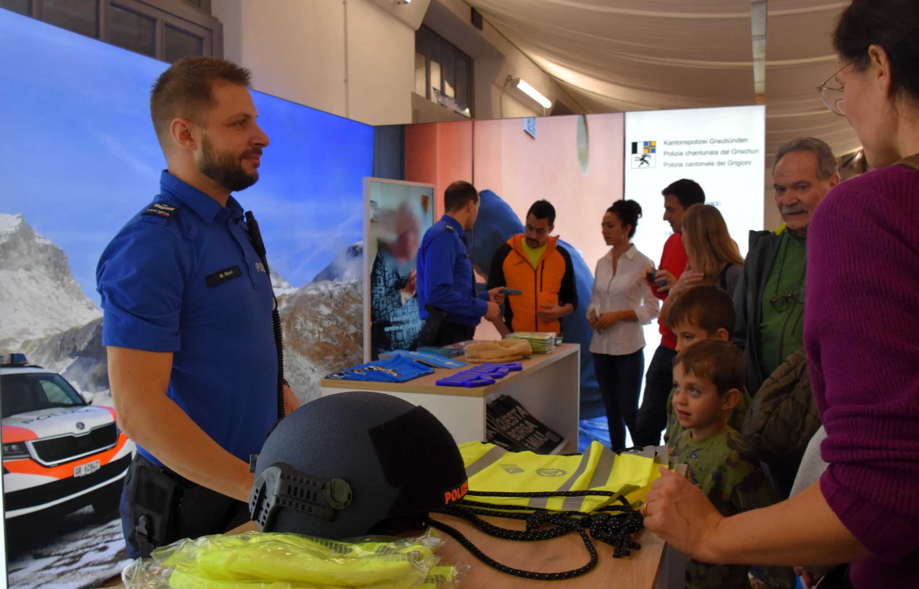 Un agente di polizia in uniforme blu interagisce con un gruppo di persone in uno stand espositivo, circondato da materiali informativi e attrezzature di sicurezza.