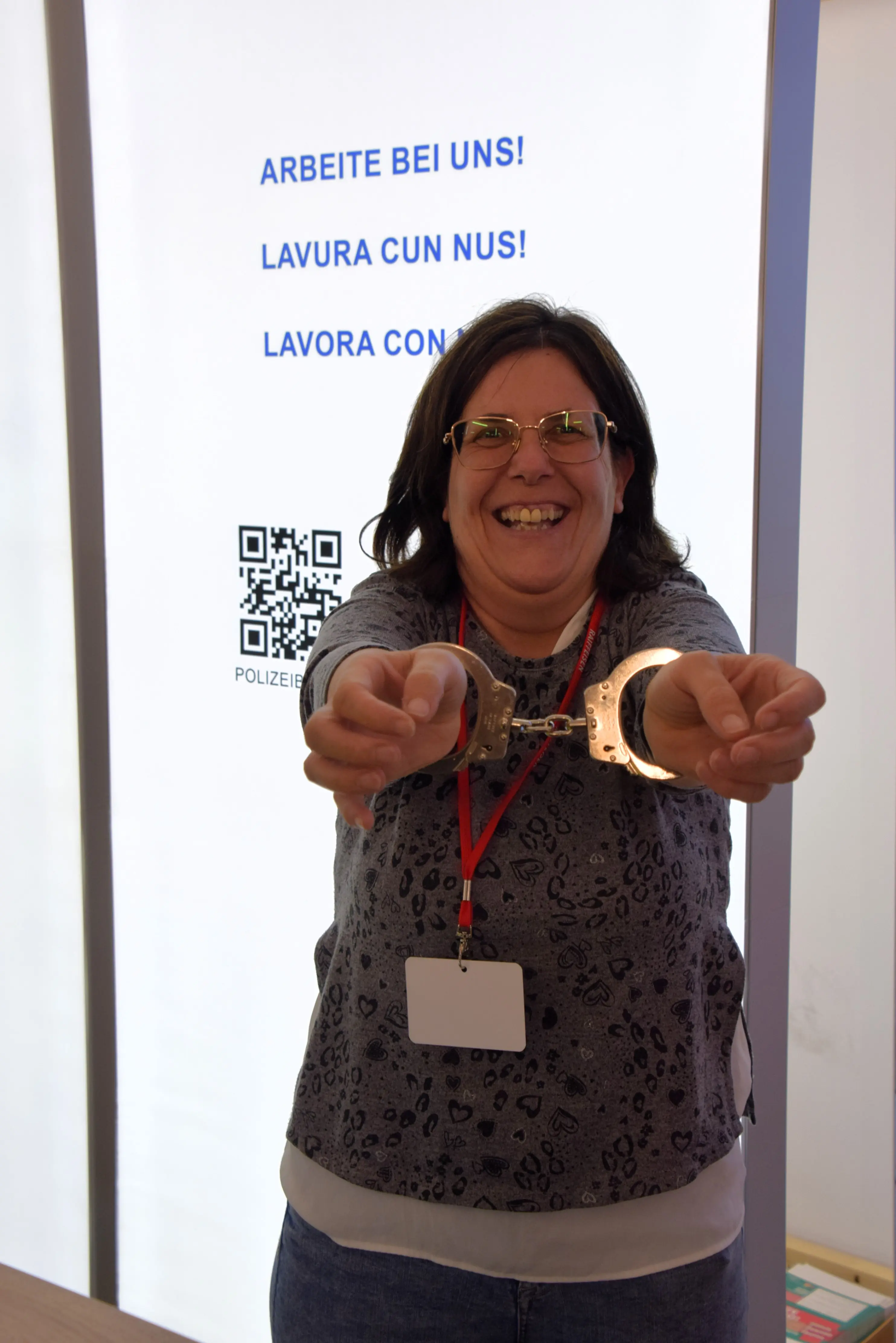 Una donna sorridente indossa manette e tiene le braccia tese in avanti. Sullo sfondo si vedono iscrizioni in tedesco e italiano che invitano a lavorare insieme.