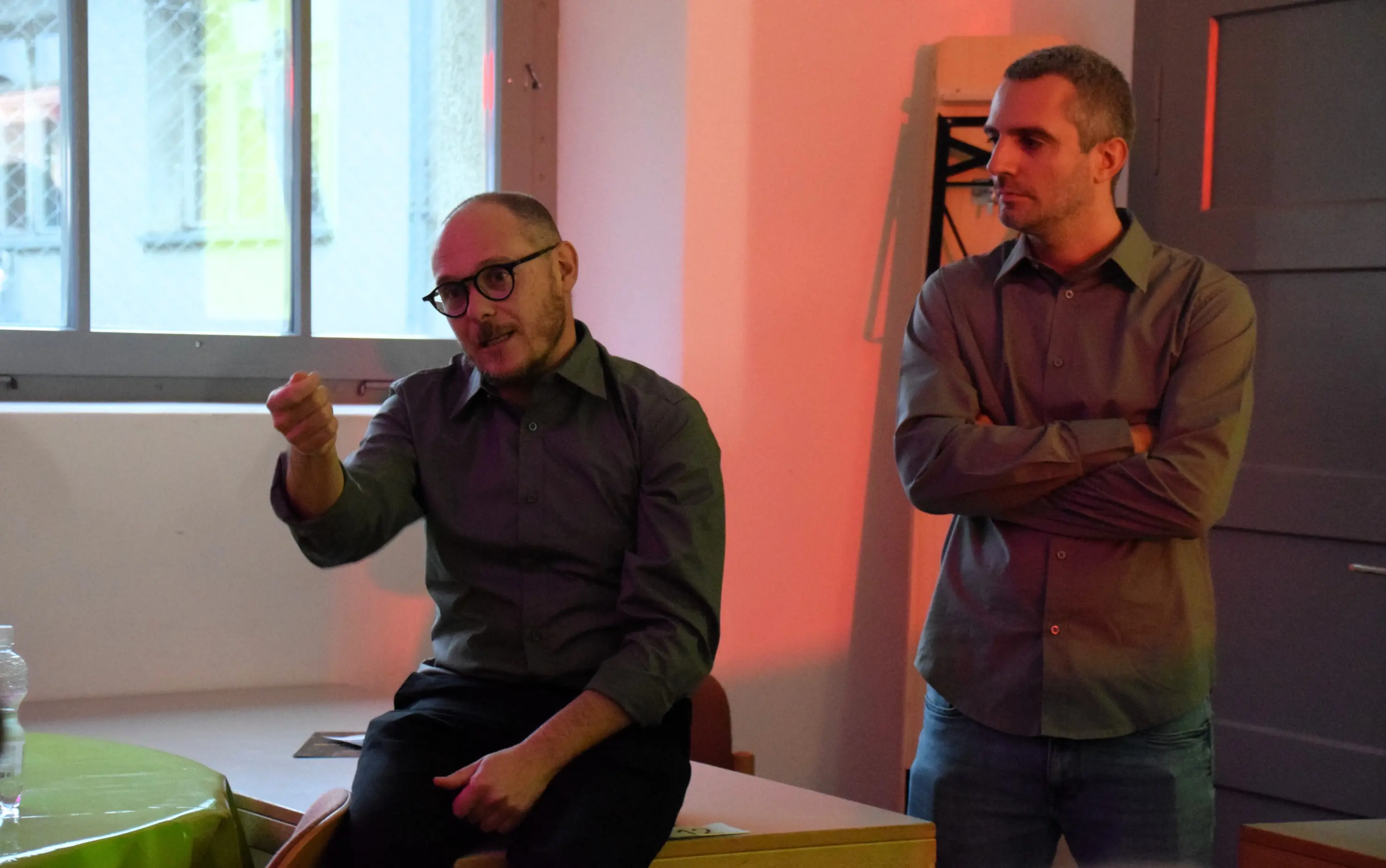 Due uomini in camicia grigia sono in un ambiente di lavoro. Uno, seduto su un tavolo, gesticola mentre parla, mentre l'altro sta in piedi accanto a lui, ascoltando attentamente. Sullo sfondo si vedono finestre e una parete