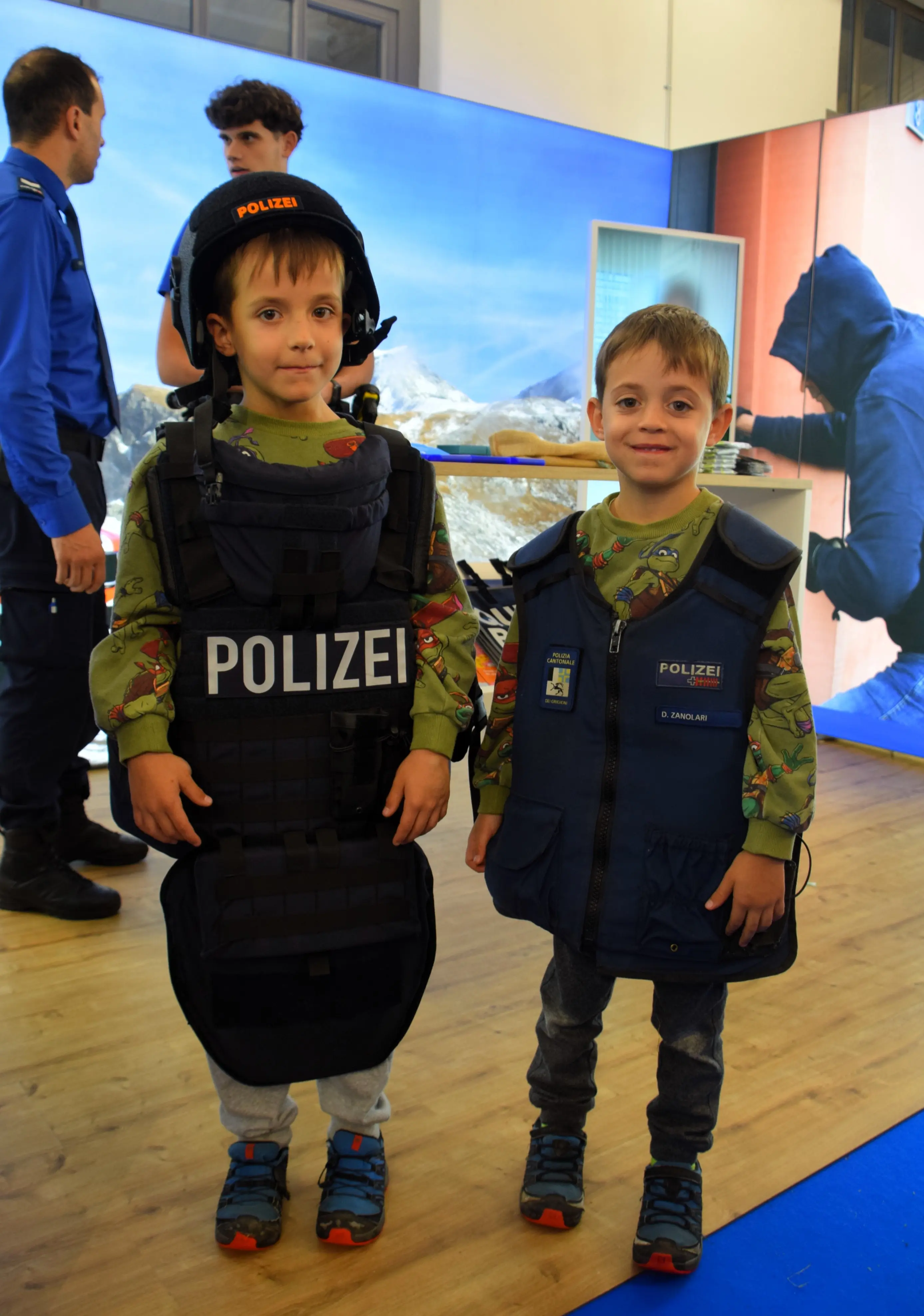 Due bambini indossano giubbotti e caschi della polizia. Sono sorridenti e si trovano in un ambiente espositivo, con adulti e interattivi sullo sfondo.