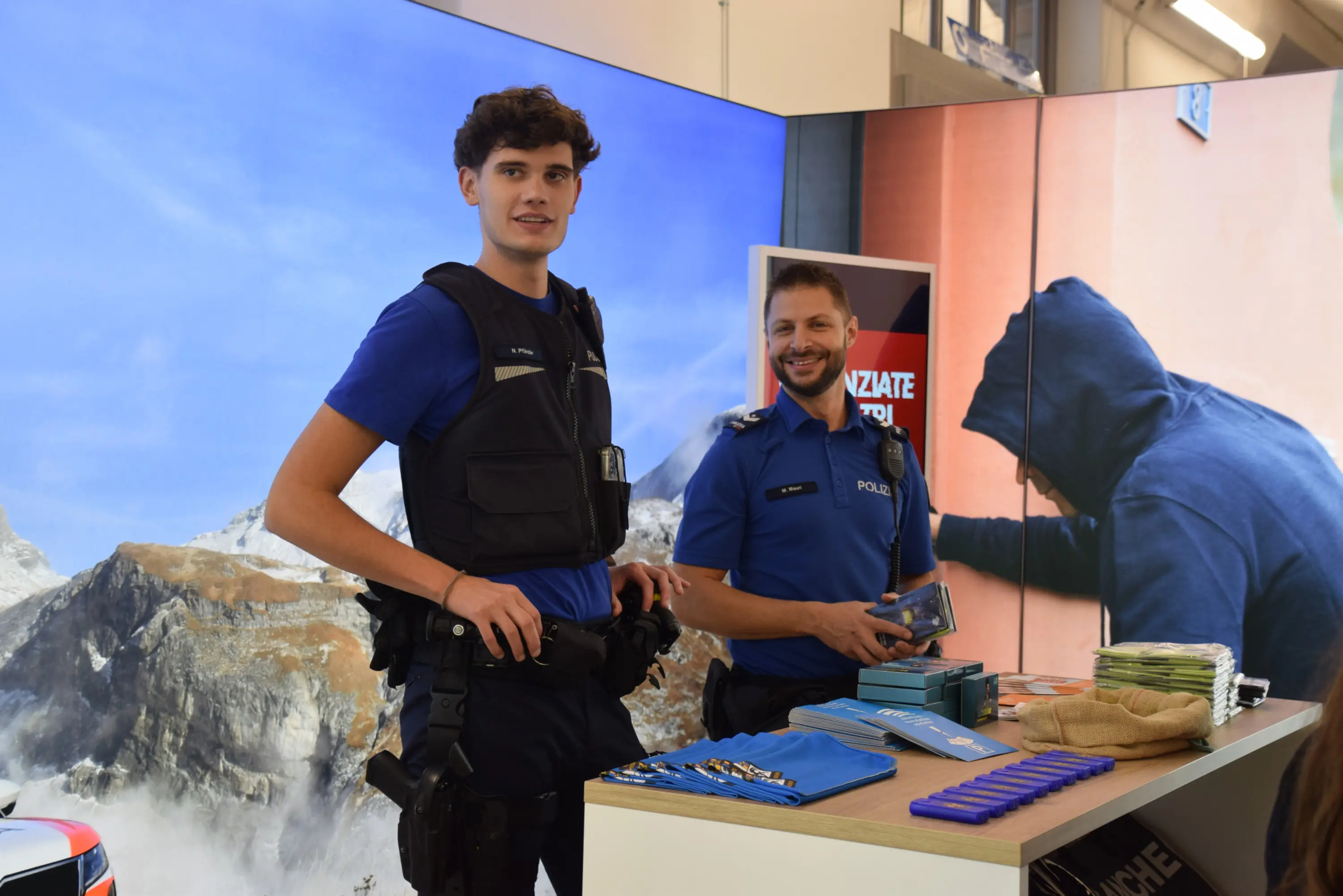 Due ufficiali di polizia sorridono dietro a un tavolo promozionale, indossando uniformi blu. Sullo sfondo si vedono montagne e una persona con cappuccio.