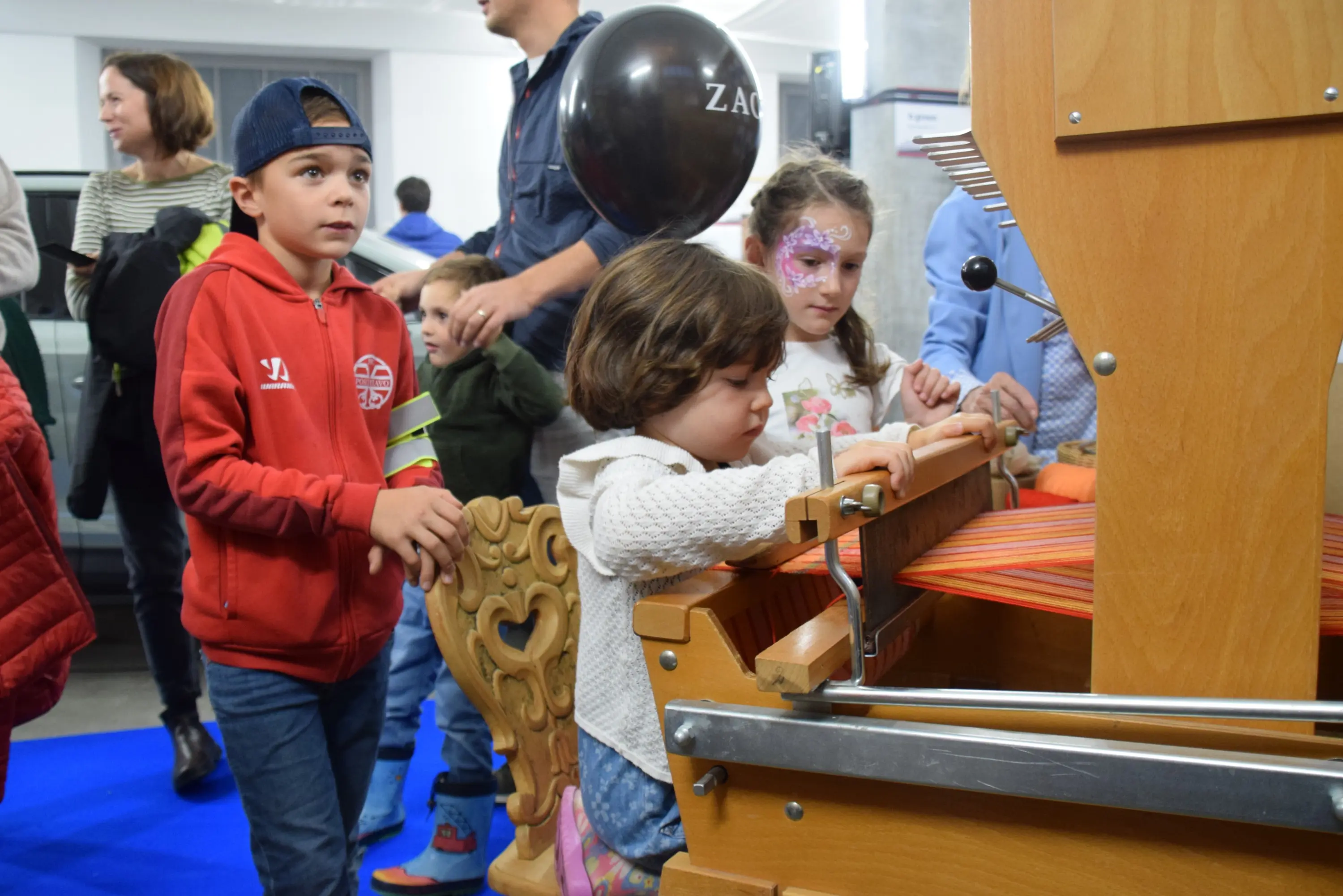 Bambini in un evento interattivo, una bambina gioca con un telaio mentre altri bambini la osservano. alcuni adulti sono in background. Una bolla nera si libra sopra la scena.