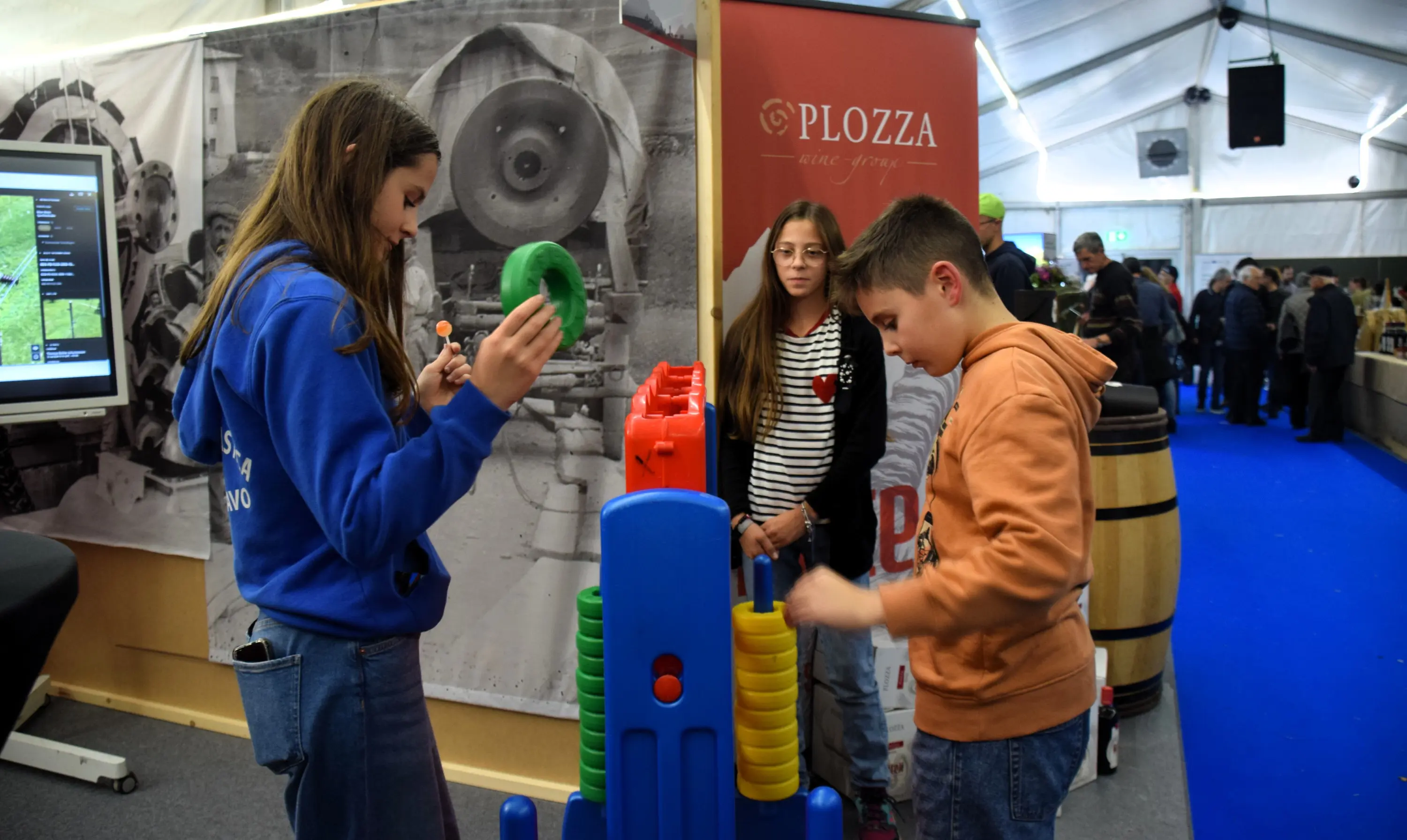 Due ragazzi giocano a un gioco educativo all'interno di una fiera. Una ragazza indossa una felpa blu e sta impilando anelli colorati, mentre un ragazzo a destra sta posizionando dei dischi su una struttura. Sullo sfondo, si ved