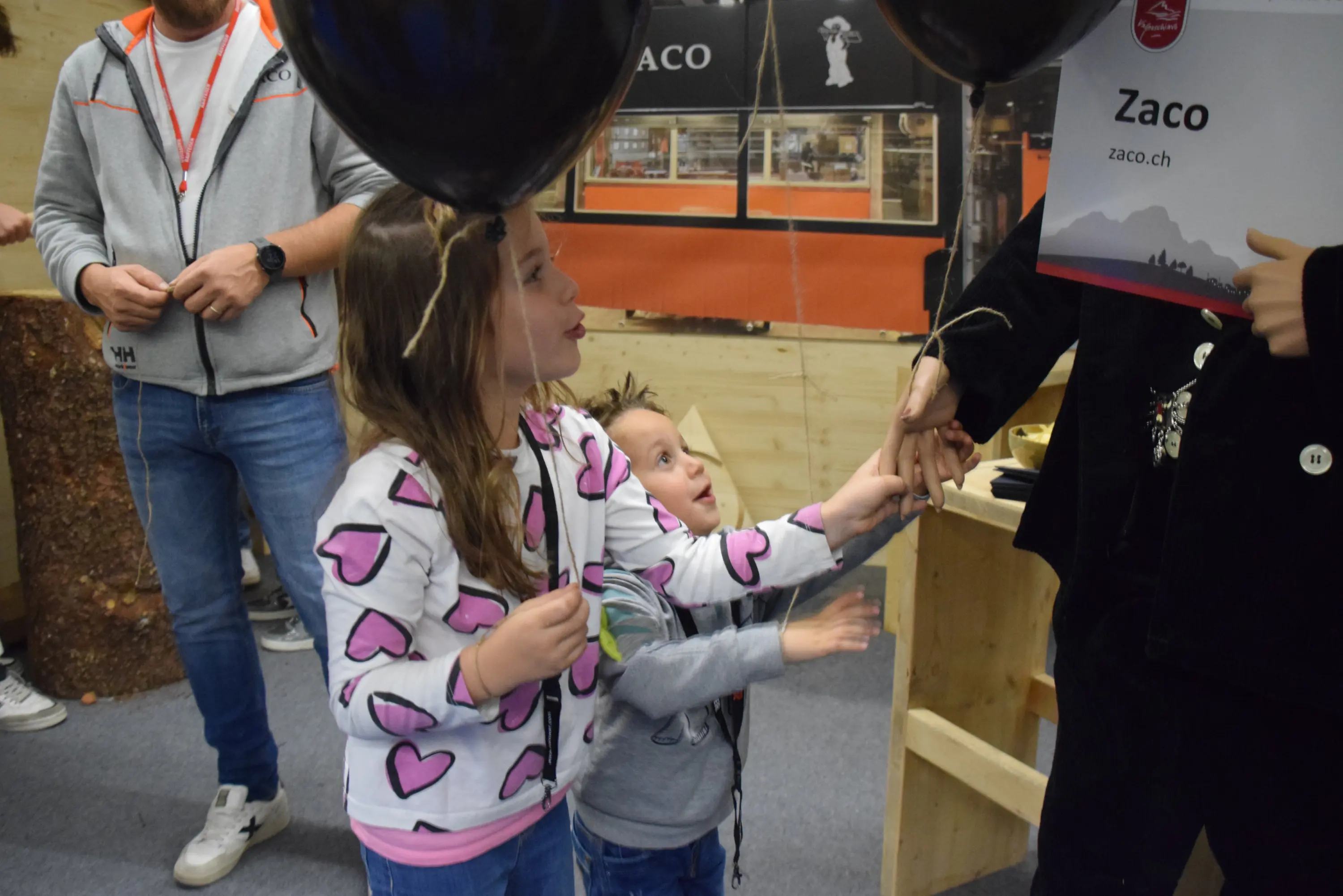 Due bambini, uno con una felpa a cuori e l'altro con una maglietta chiara, guardano incuriositi un adulto che tiene un cartello con il nome "Zaco" e dei palloncini neri. Sullo sfondo si vedono elementi di leg