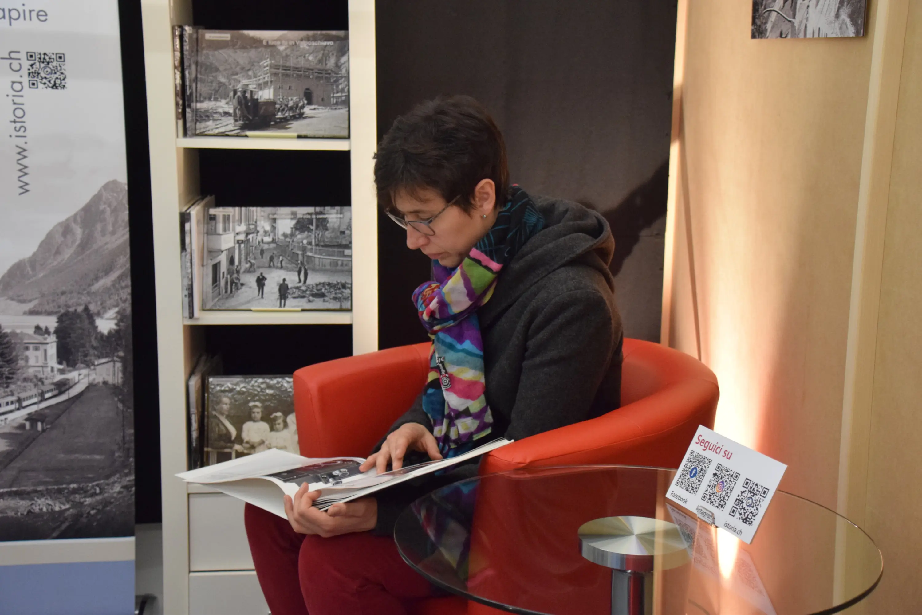 Una donna seduta su una sedia rossa legge un libro fotografico, mentre si trova in un ambiente espositivo con immagini storiche sullo sfondo.