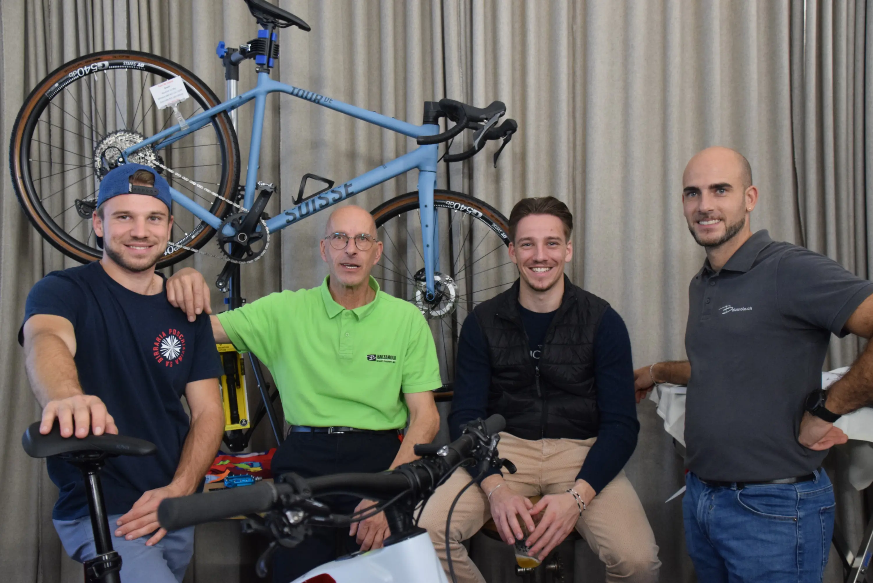 Quattro uomini sorridenti posano insieme davanti a delle biciclette esposte. Due di loro sono seduti, mentre gli altri due stanno in piedi, tutti indossano abbigliamento casual.