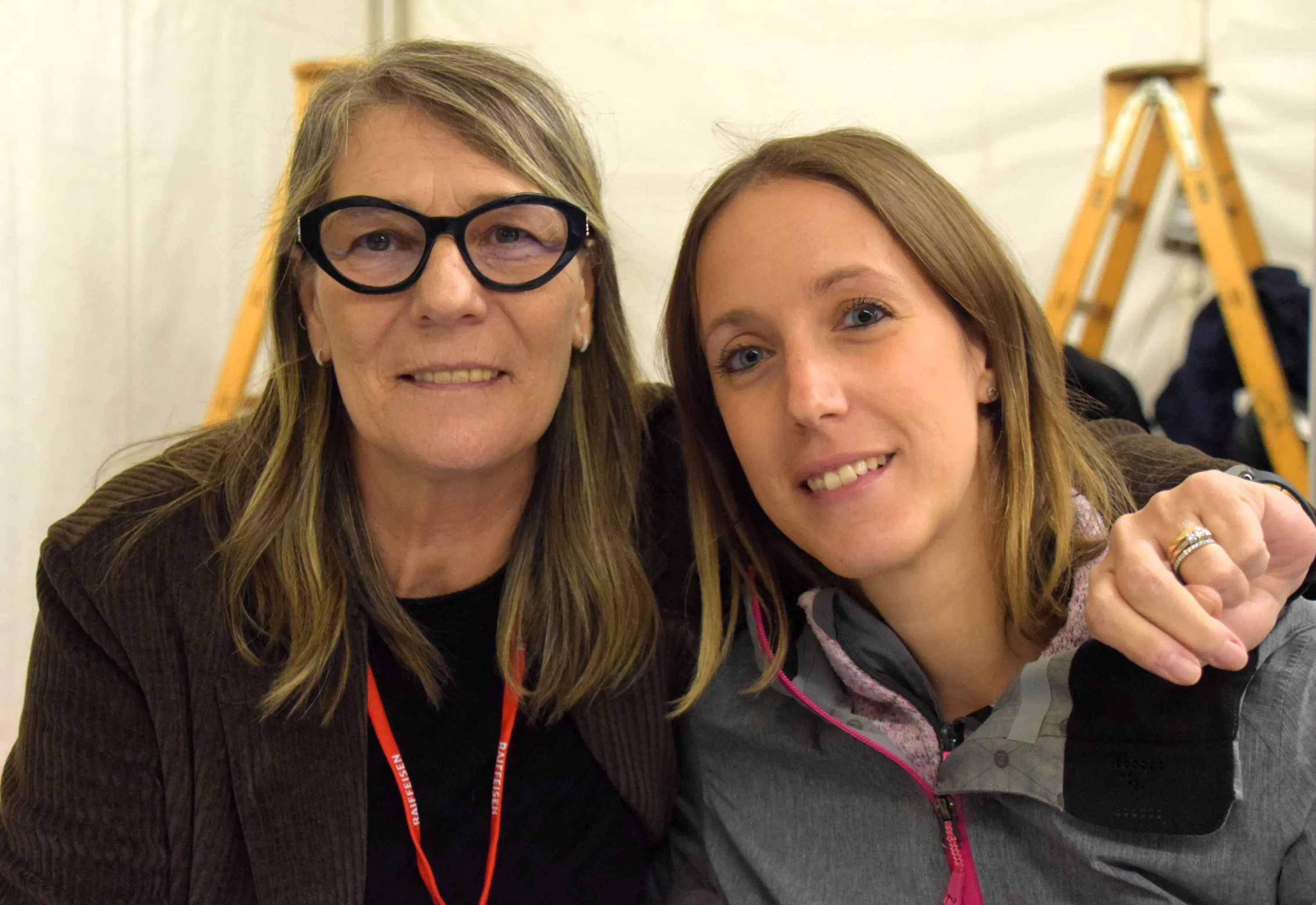 Due donne sorridenti si trovano sedute insieme in un ambiente accogliente. La donna a sinistra ha i capelli lunghi e indossa occhiali neri, mentre quella a destra ha i capelli più corti e indossa una giacca grigia.