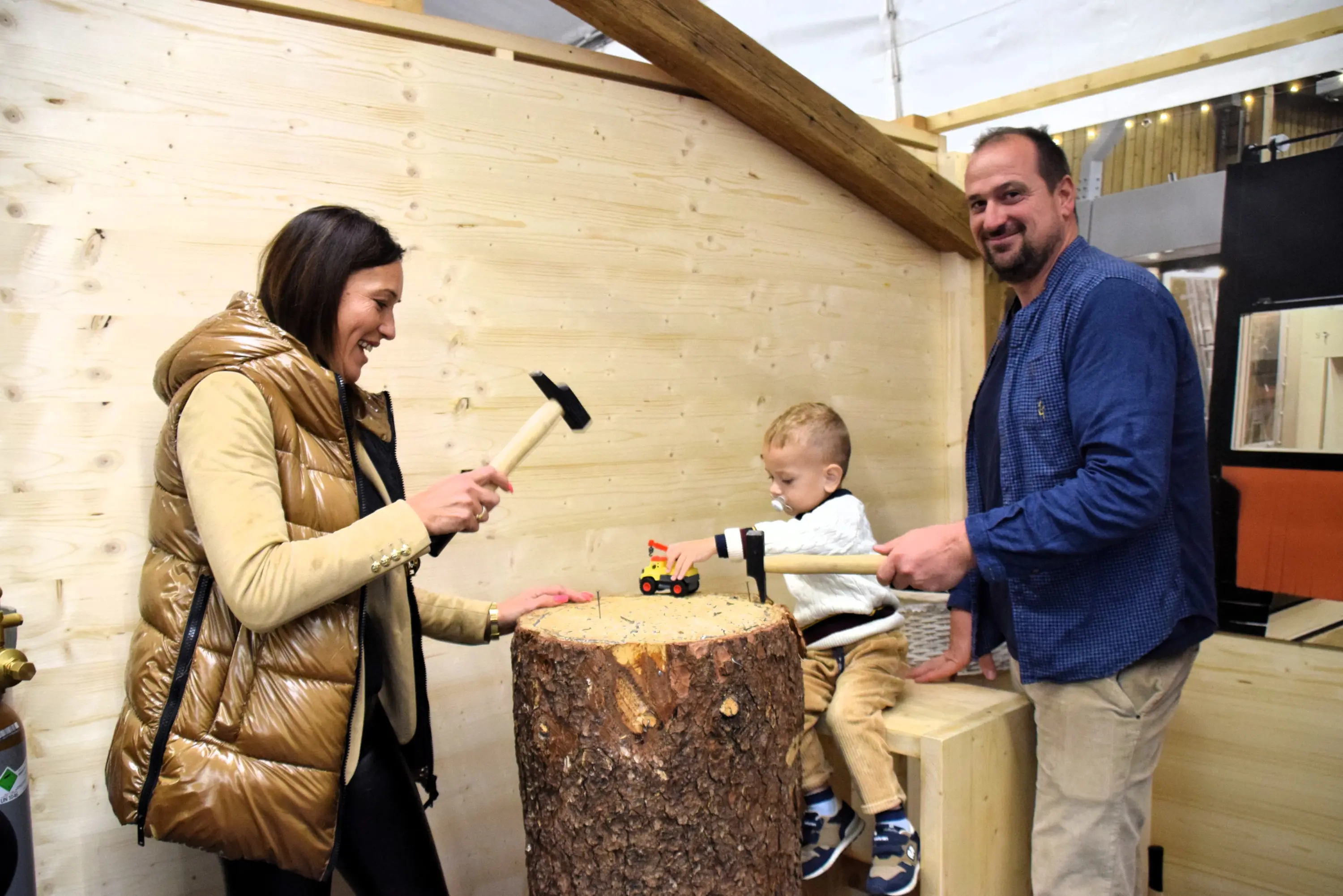 Una donna col martello e un uomo sono sorridenti mentre giocano con un bambino su un ceppo di legno. Il bambino gioca con un giocattolo a forma di trattore. Il fondo è una struttura di legno.