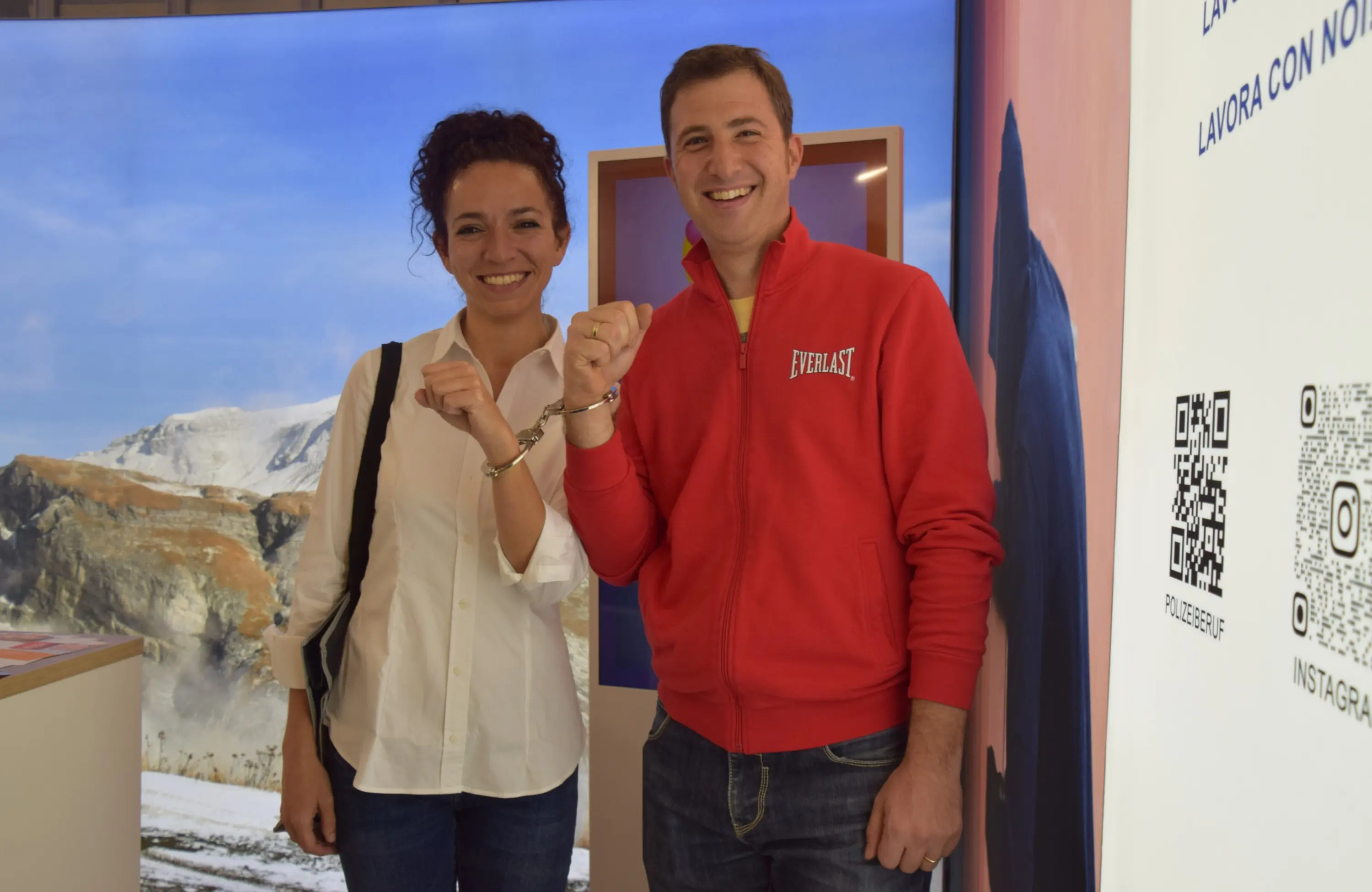 Due persone sorridenti fanno un gesto con le mani, mostrando i polsi uniti, sullo sfondo di un paesaggio montano e una parete colorata.