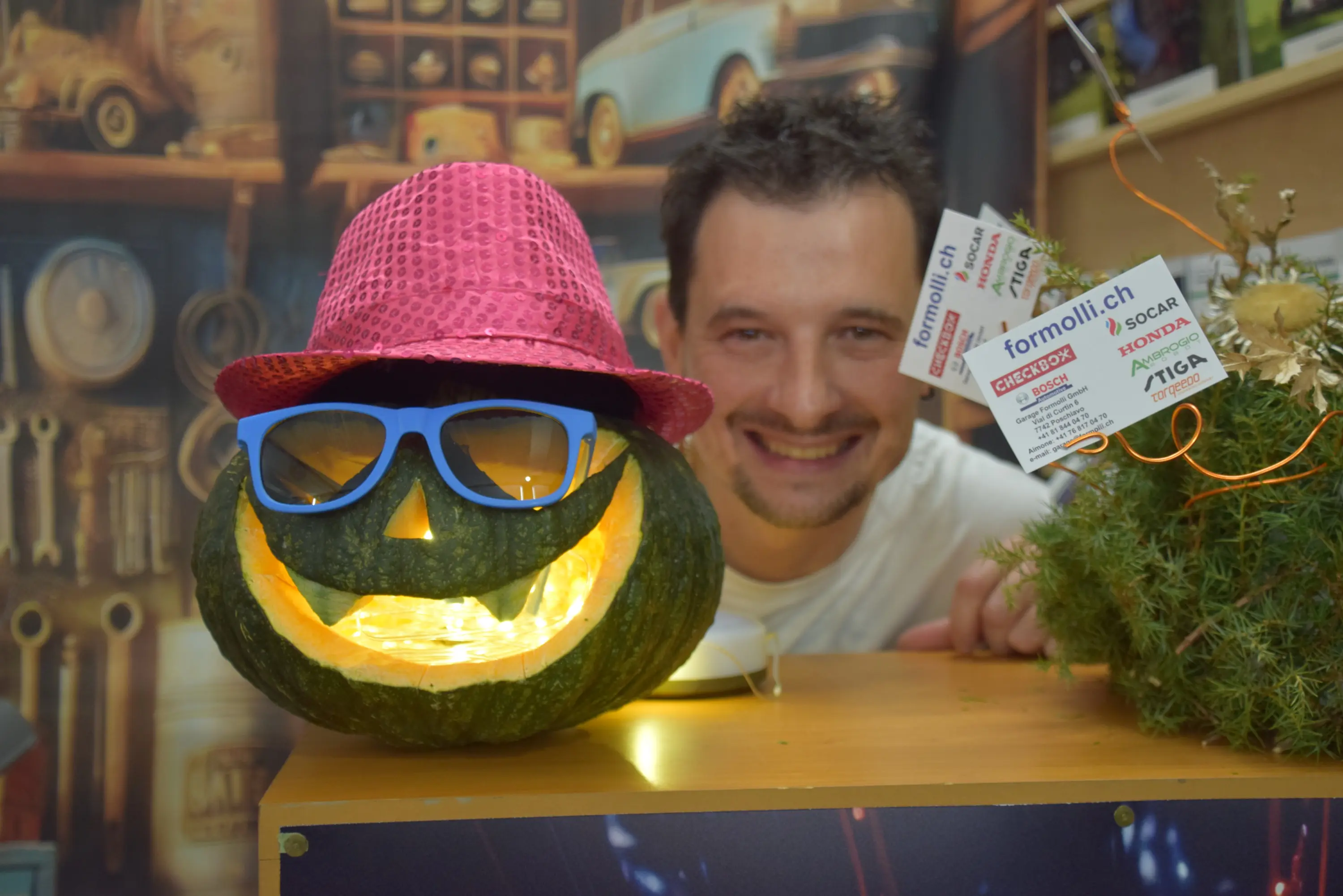 Un uomo sorridente si trova dietro a una zucca intagliata con un sorriso, adornata con occhiali da sole e un cappello rosa. Sul tavolo ci sono biglietti e decorazioni autunnali.