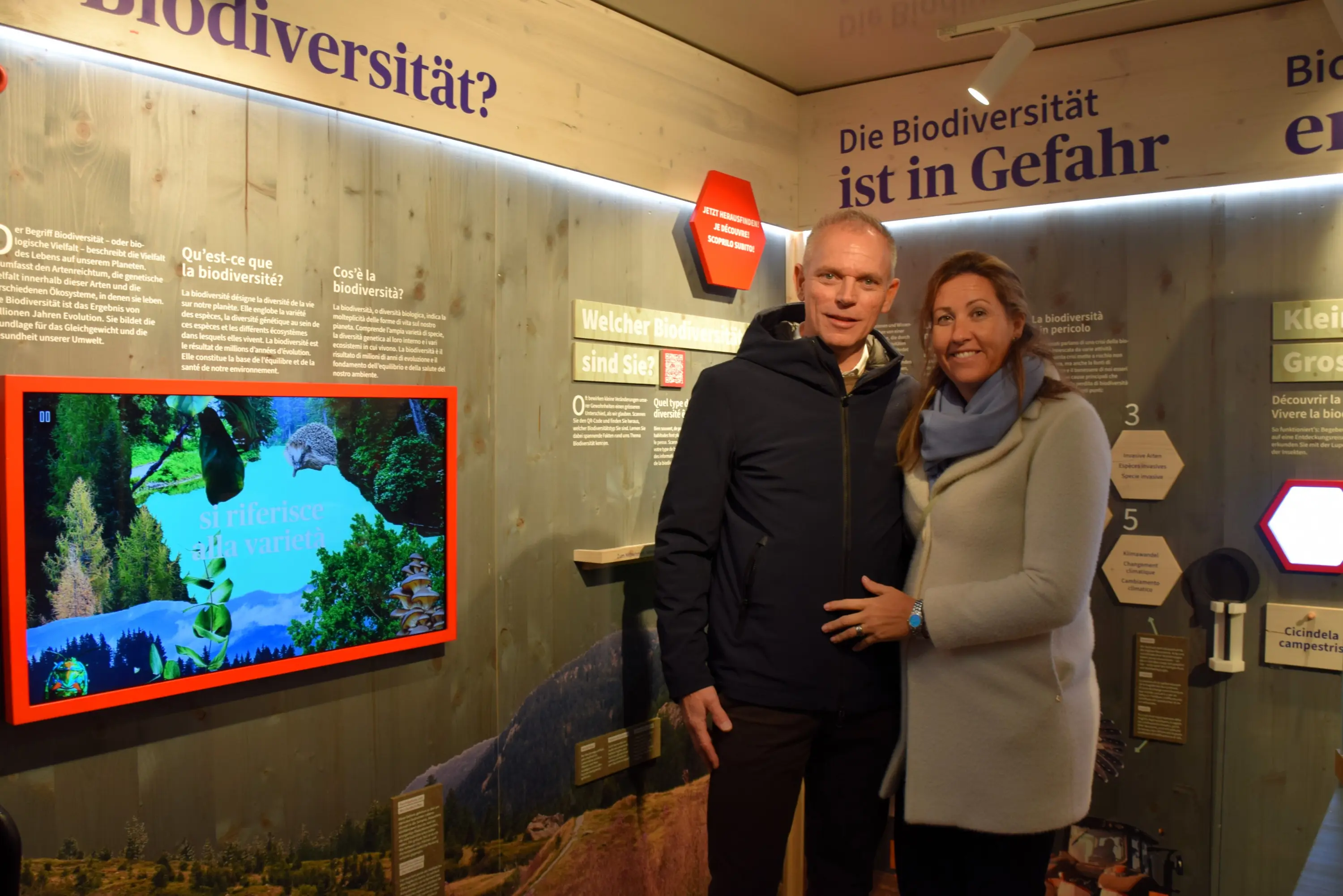 Una coppia sorridente si trova in un museo, di fronte a un pannello informativo sulla biodiversità. Sullo sfondo, è visibile un video che mostra paesaggi naturali e informazioni sulla varietà ecologica.