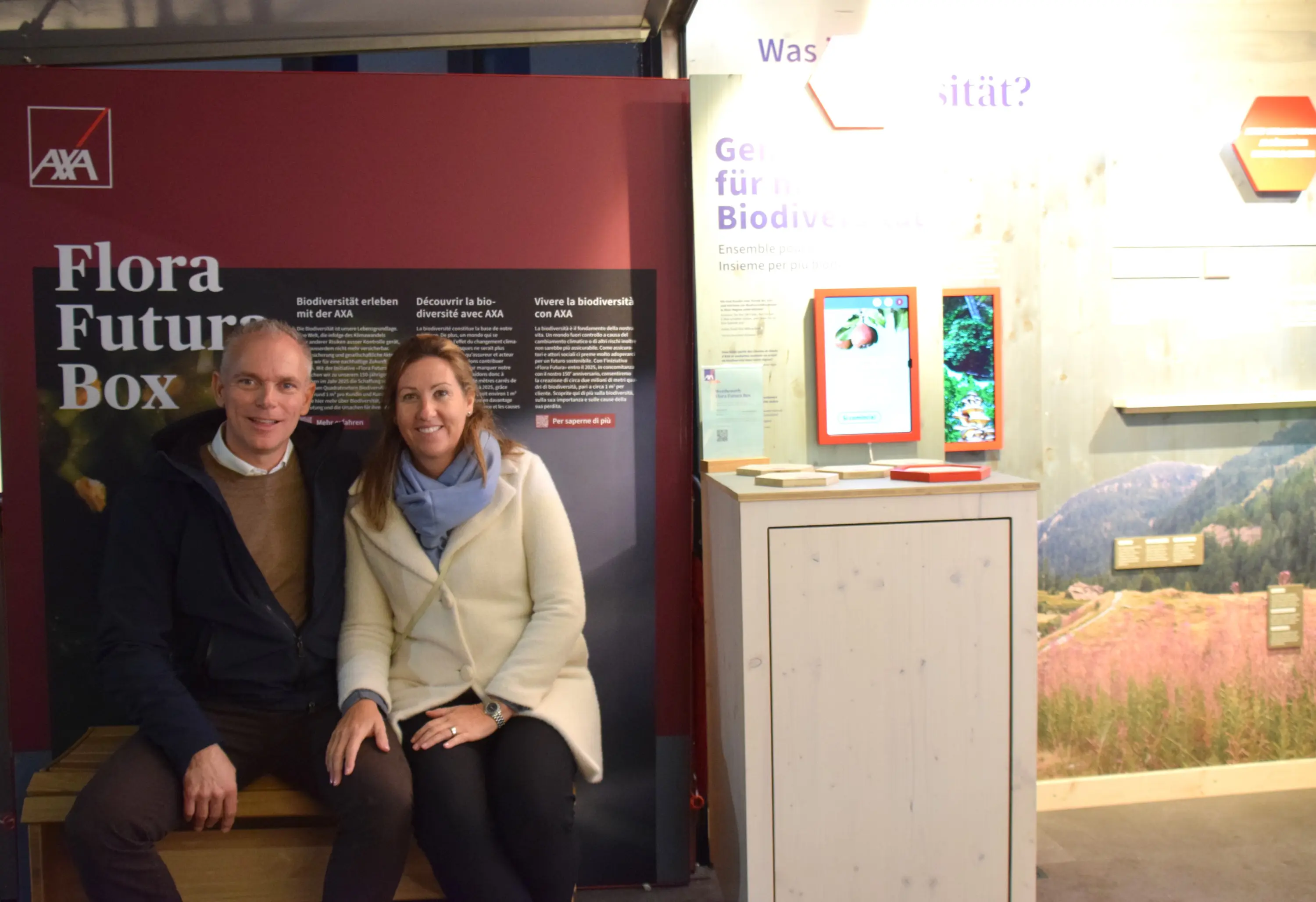 Due persone sorridenti sedute su una panchina davanti a un'esposizione informativa sulla biodiversità. Sullo sfondo, si vedono pannelli con testo esplicativo e immagini della natura.