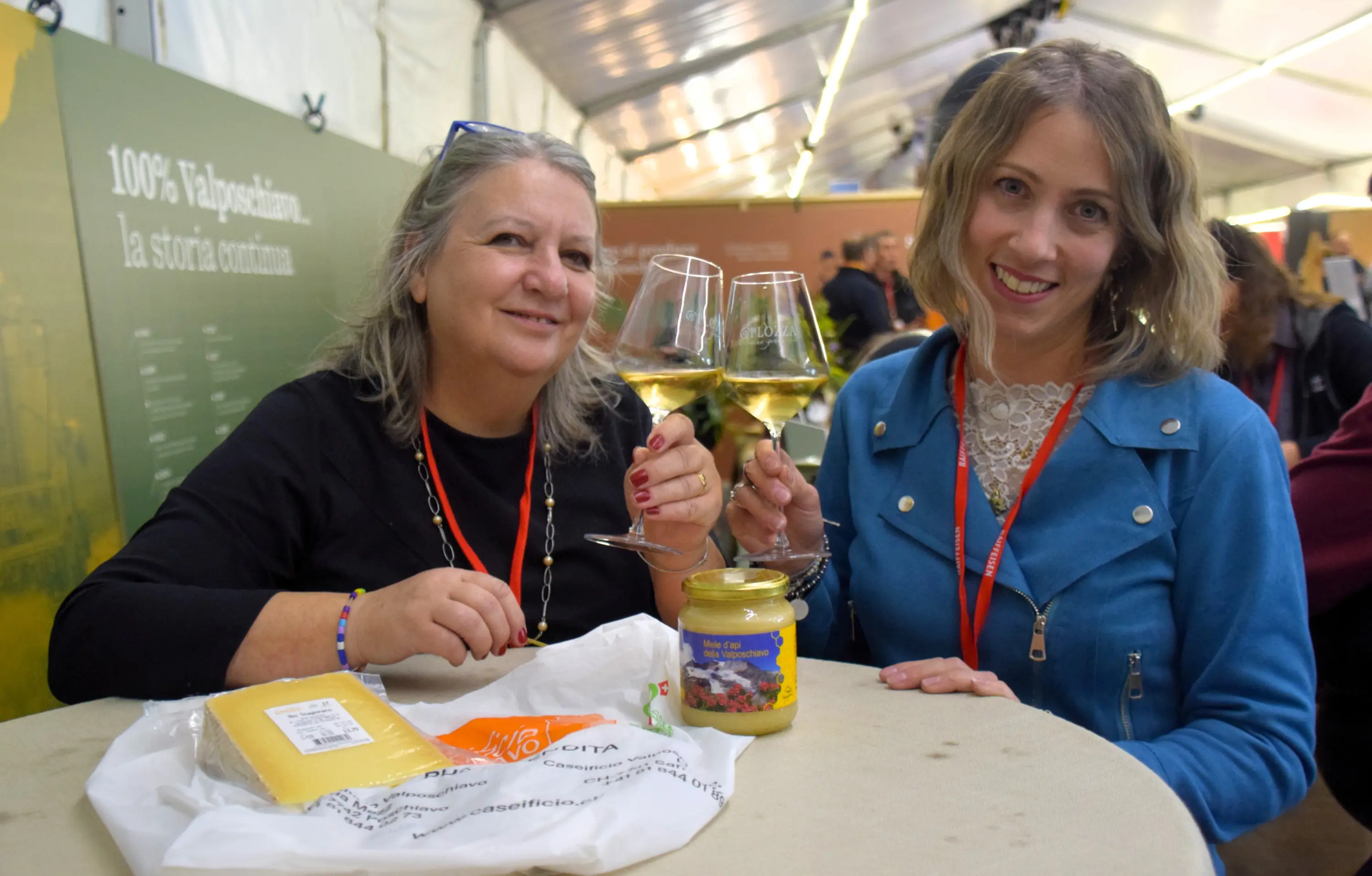 Due donne sorridenti brindano con bicchieri di vino bianco, sedute a un tavolo. Sul tavolo ci sono un barattolo di marmellata e un sacchetto di formaggio. Dietro di loro si intravede un evento affollato.