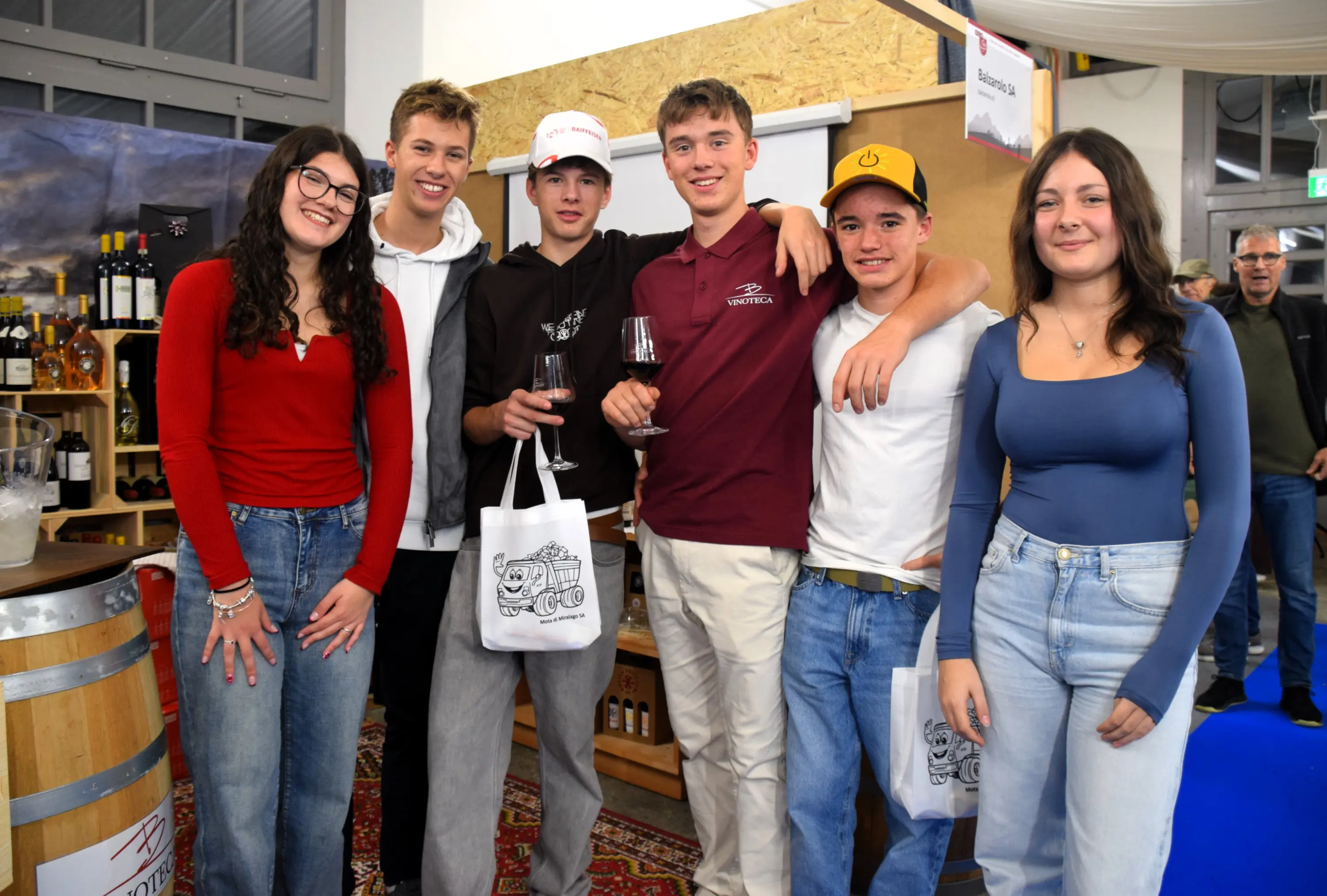 Gruppo di sei giovani sorridenti che posano insieme in un ambiente di degustazione di vini. Alcuni tengono in mano un bicchiere di vino e altre borse di tela. Sullo sfondo si vedono botti di legno e scaffali con bottiglie di vino.