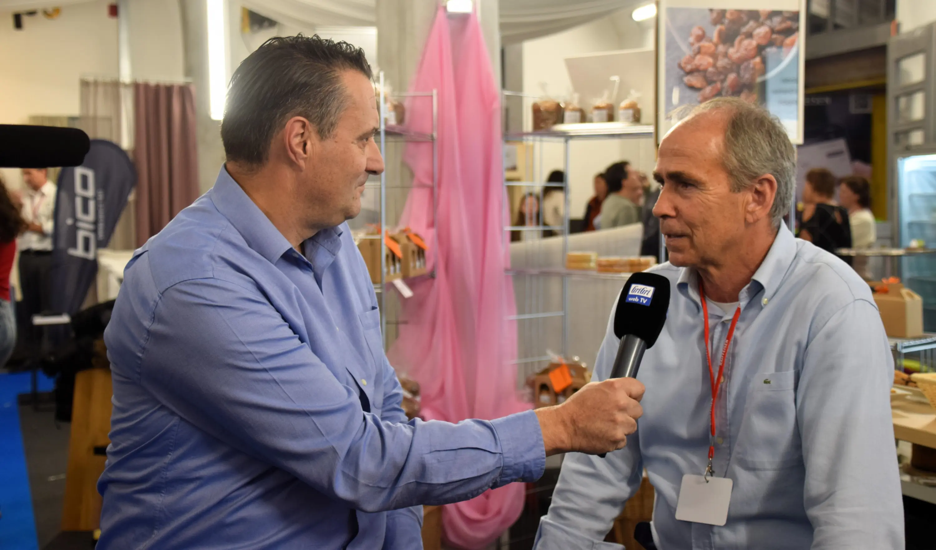 Due uomini stanno conversando a un evento, uno dei quali tiene un microfono e fa un'intervista. Sullo sfondo si vedono prodotti esposti e un'atmosfera di fiera.