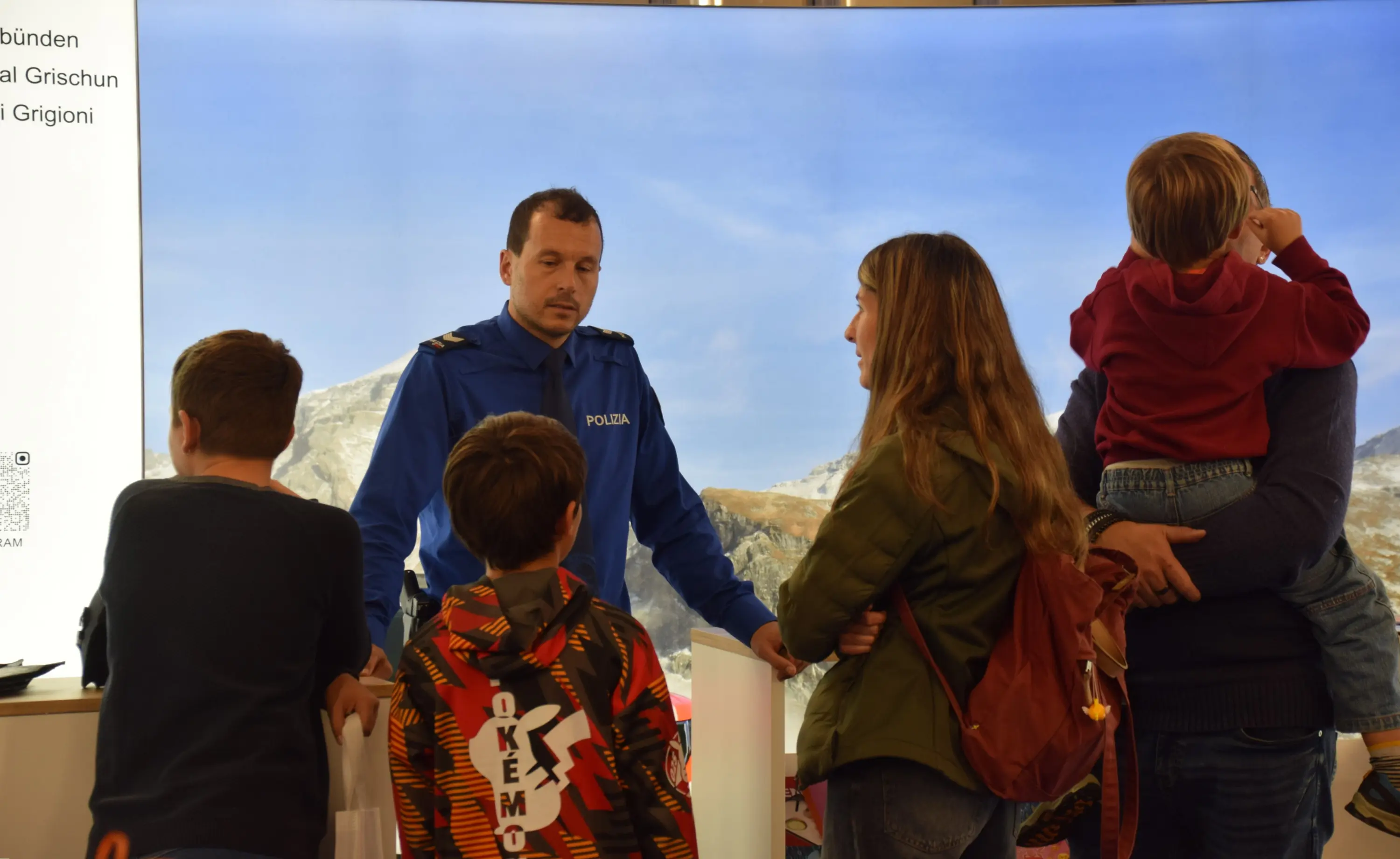 Un agente di polizia parla con un gruppo di persone, tra cui bambini, in un ambiente informativo con uno sfondo montano.