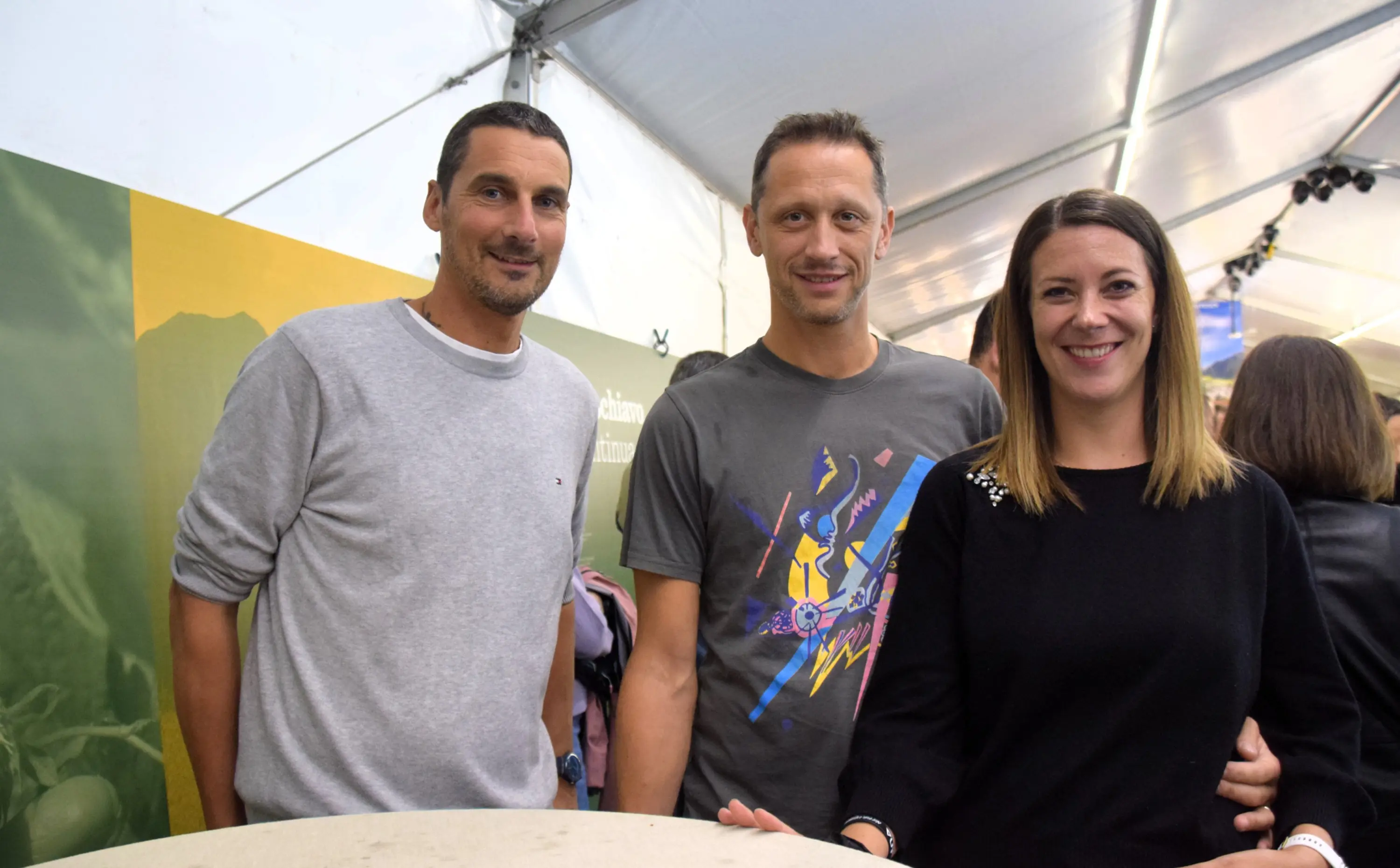 Tre persone sorridenti in un ambiente all'aperto, all'interno di una tenda. Due uomini e una donna, abbigliamento informale, posano insieme con un'atmosfera amichevole. Sullo sfondo, una parete colorata con scritte.