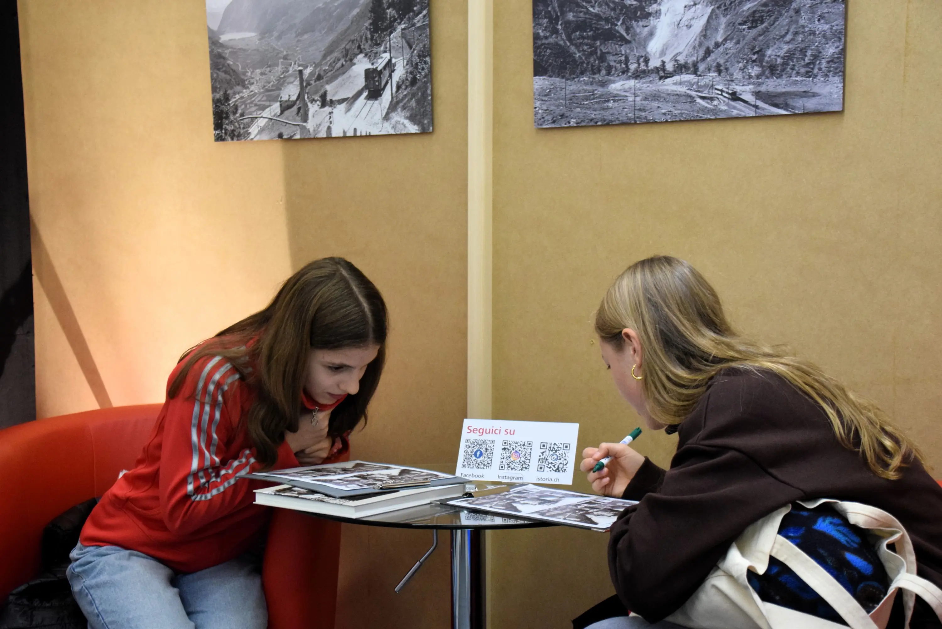 Due ragazze si trovano a un tavolo. Una di loro indossa una felpa rossa e si inclina in avanti, mentre l'altra scrive su un foglio. Sul tavolo ci sono fotografie in bianco e nero e un cartello con un codice QR. Sullo