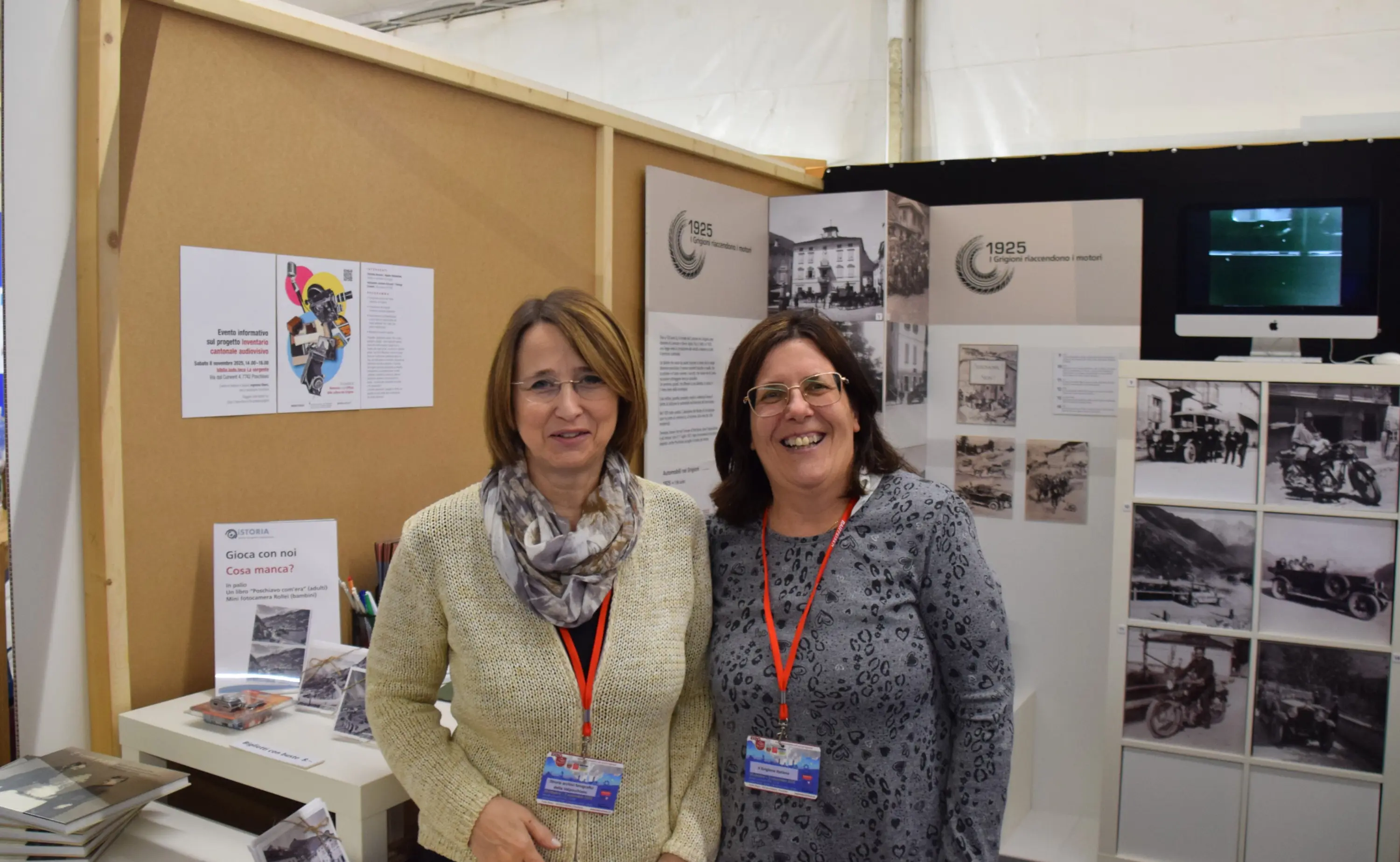 Due donne sorridenti posano davanti a uno stand informativo con foto storiche e materiale promozionale. Entrambe indossano un badge e una sciarpa grigia. Sullo sfondo si vedono pannelli con testi e immagini in bianco e nero.