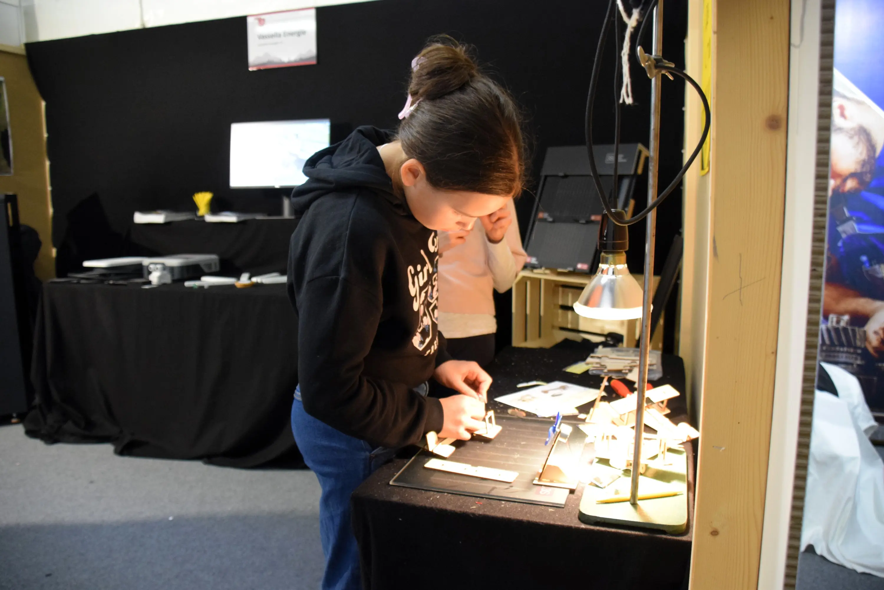 Una ragazza sta lavorando a un progetto su un tavolo illuminato, con strumenti e materiali disposti ordinatamente. Sullo sfondo si vedono altre persone e attrezzature.