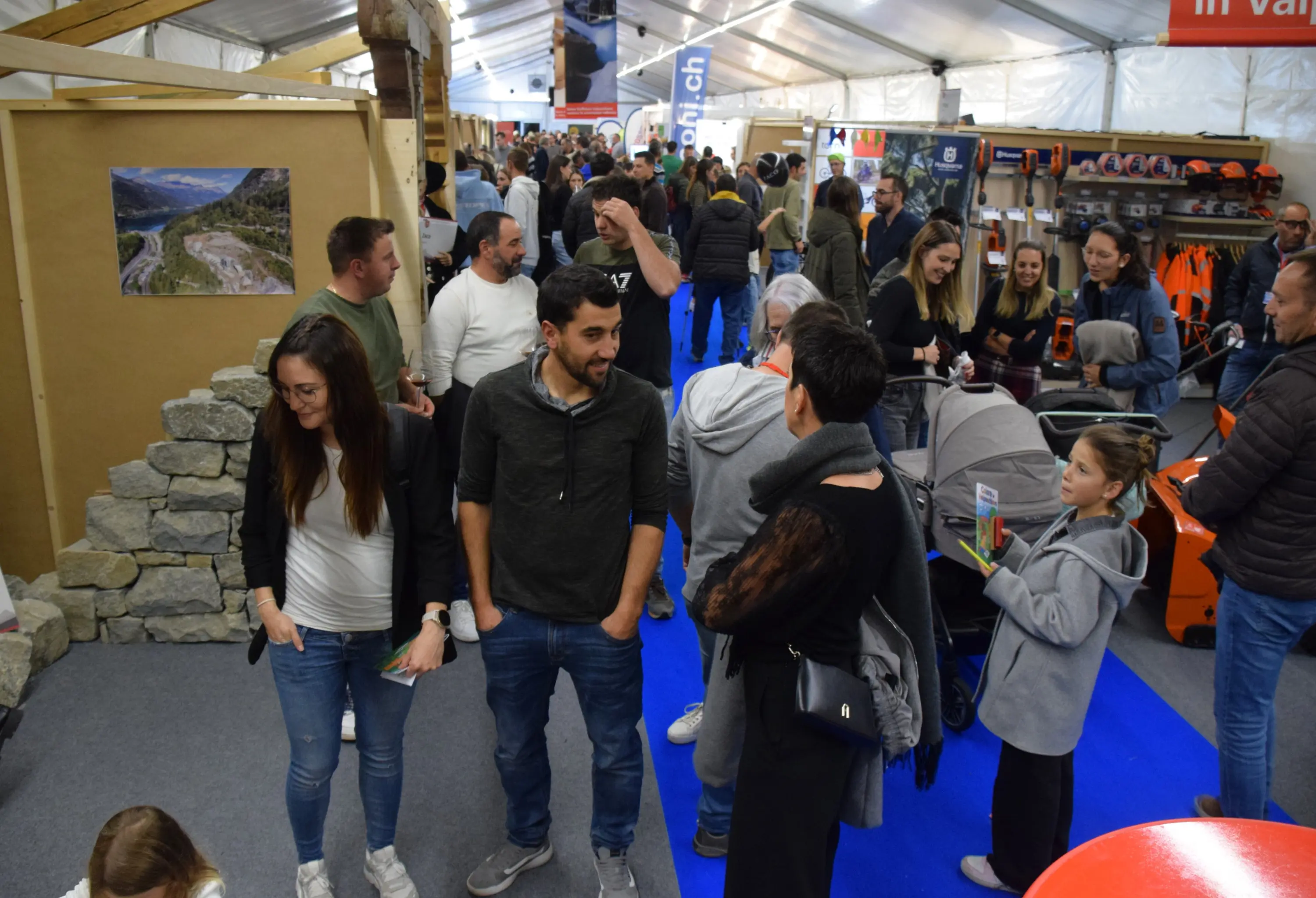 Una fiera affollata con diverse persone che interagiscono tra loro. Si vedono bambini in passeggini e stand espositivi sullo sfondo. La scena è vivace e colorata.