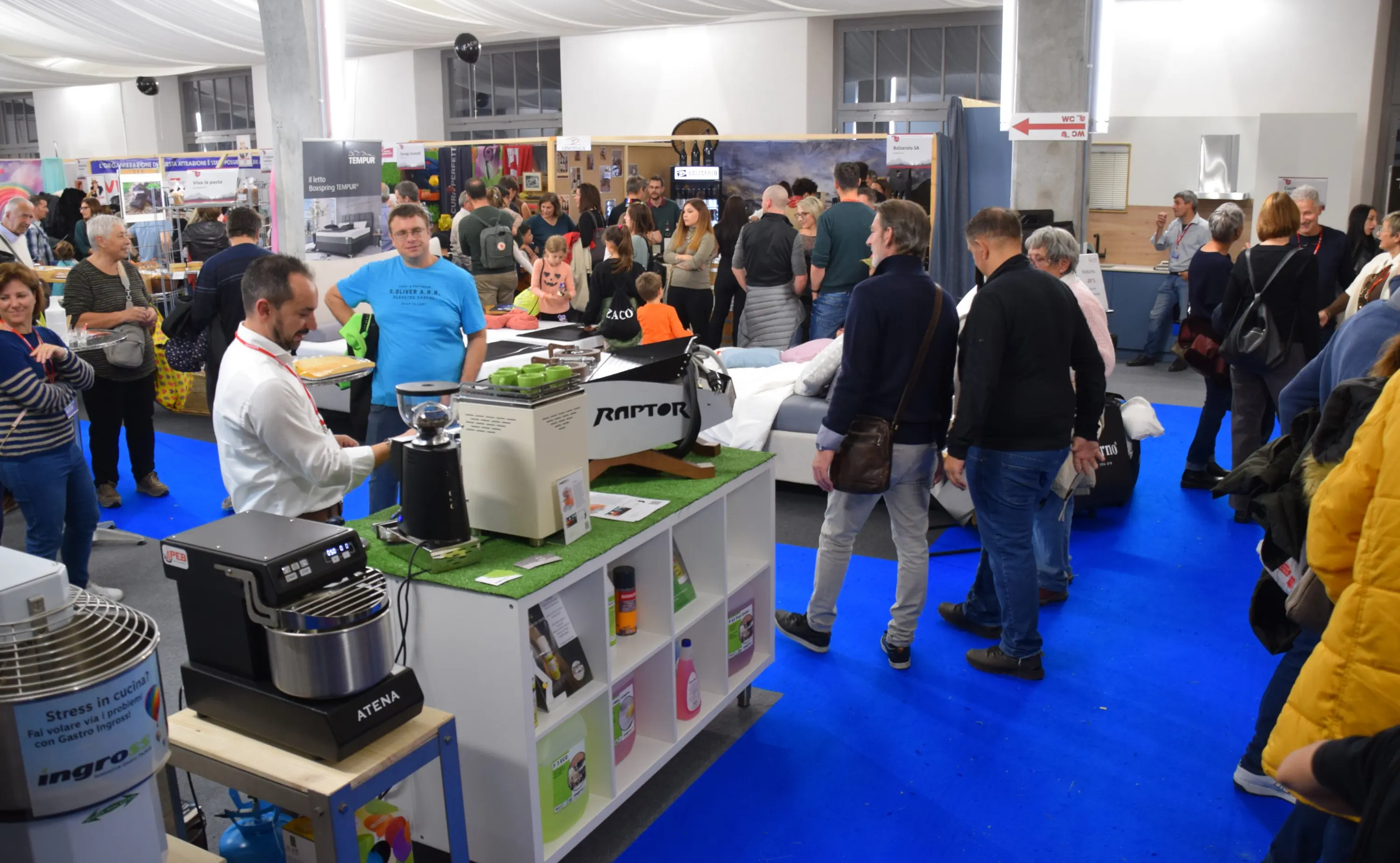 Folla di visitatori che esplorano uno spazio espositivo, con vari stand e dimostrazioni di prodotti. Sul fronte, un banco espositivo con una macchina da caffè e vari articoli in mostra.