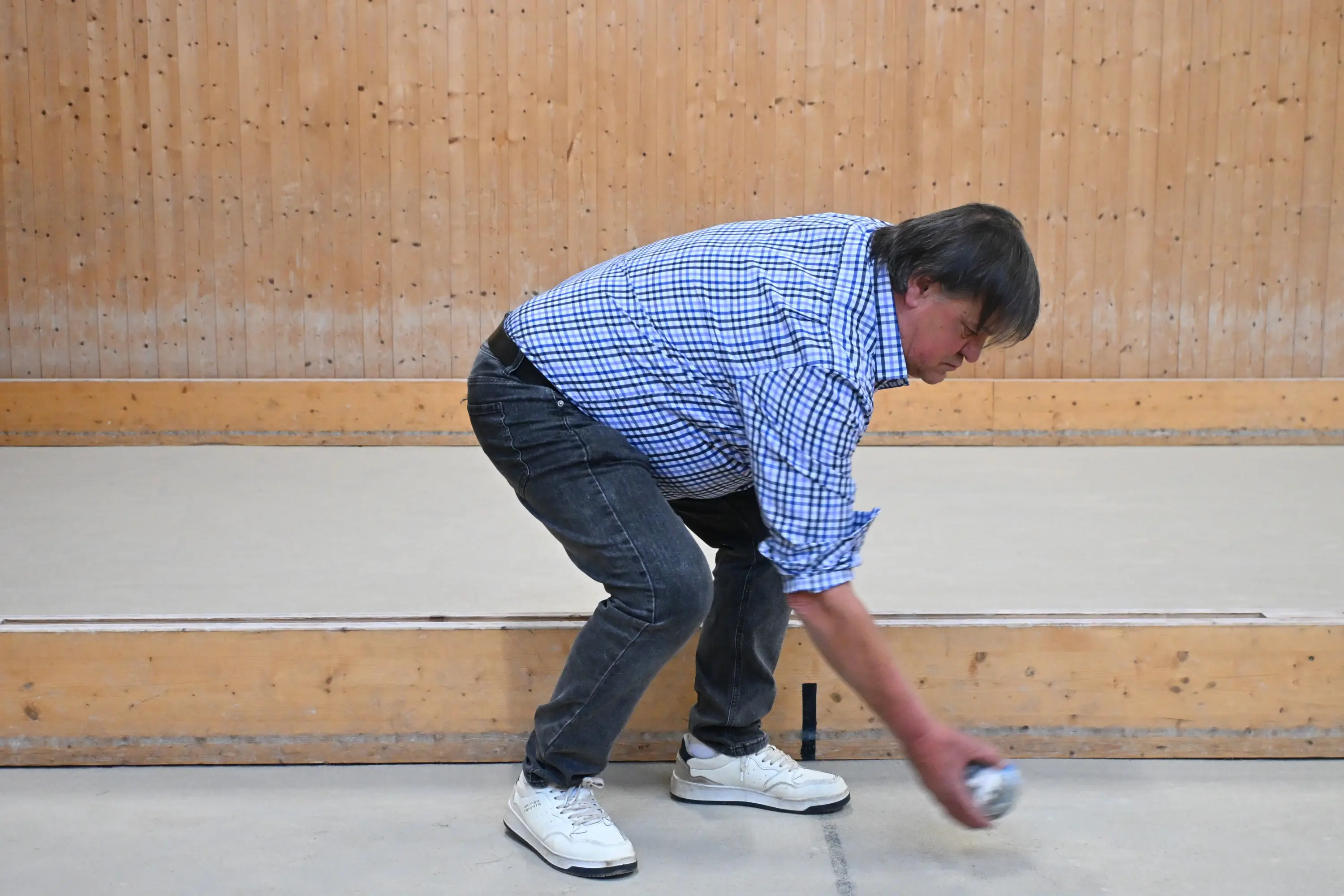 Uomo con camicia a quadri e jeans, che si piega per lanciare una boccia su un pavimento in legno, in un ambiente interno.