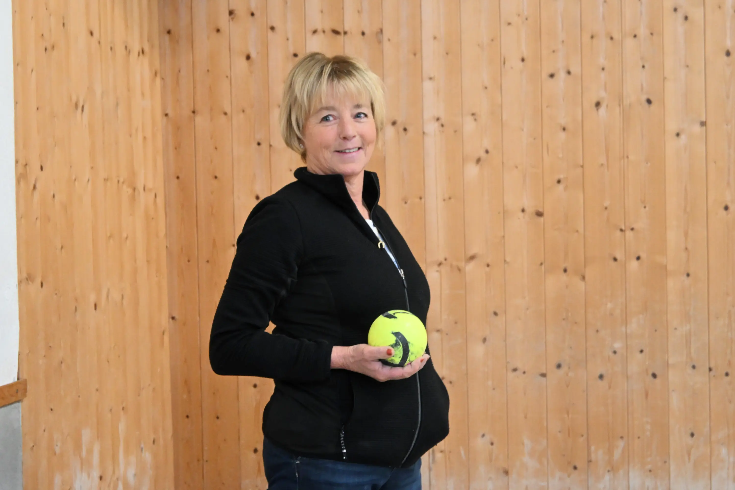 Donna sorridente con capelli corti, che indossa una giacca nera, tiene in mano una palla gialla su sfondo di muro di legno chiaro.