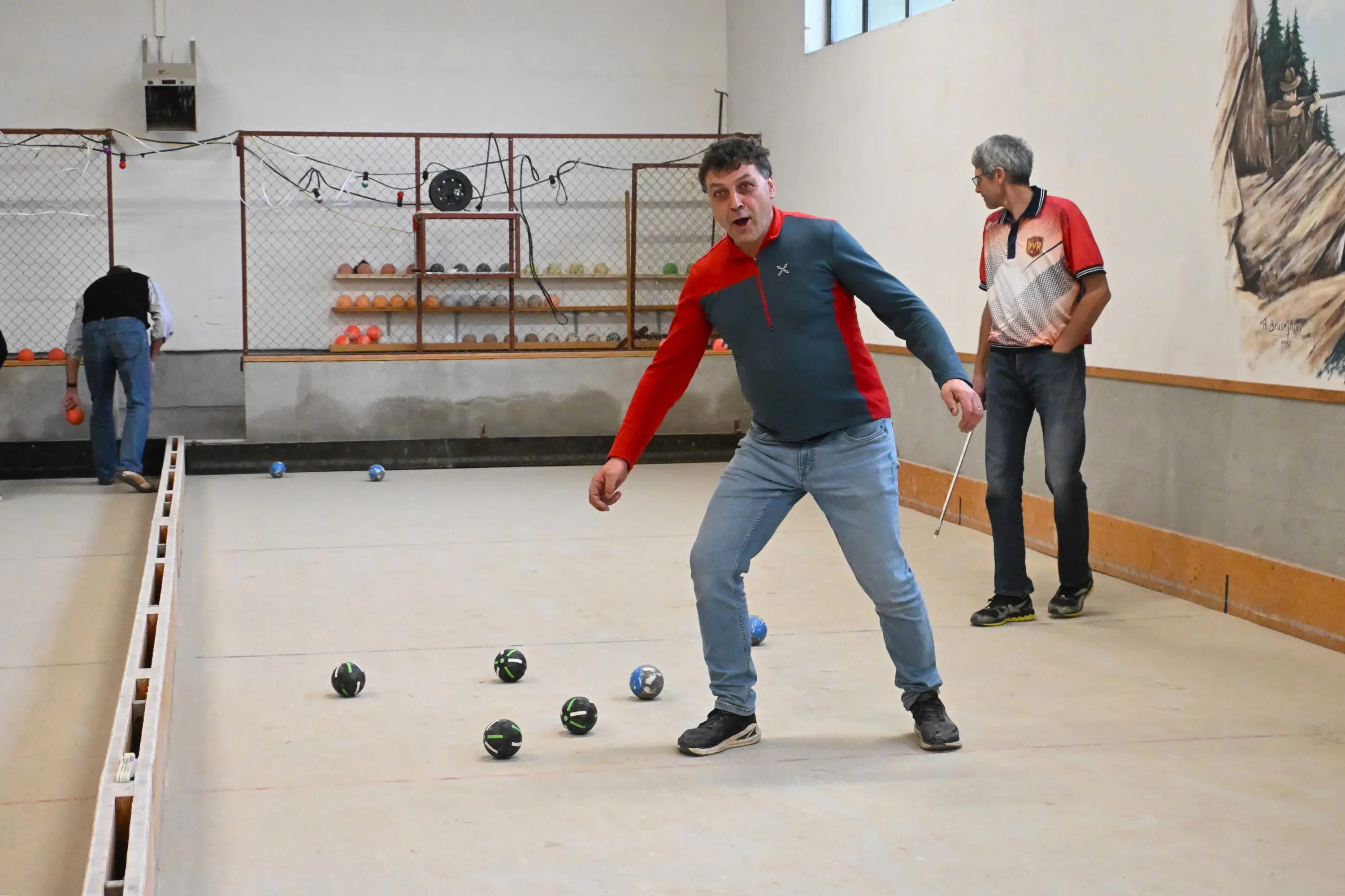 Due uomini giocano a bocce in una pista interna. Un uomo, in maglia grigia e rossa, si prepara a lanciare una boccia, mentre l'altro osserva. Sullo sfondo si vedono altri pallini disposti ordinatamente.